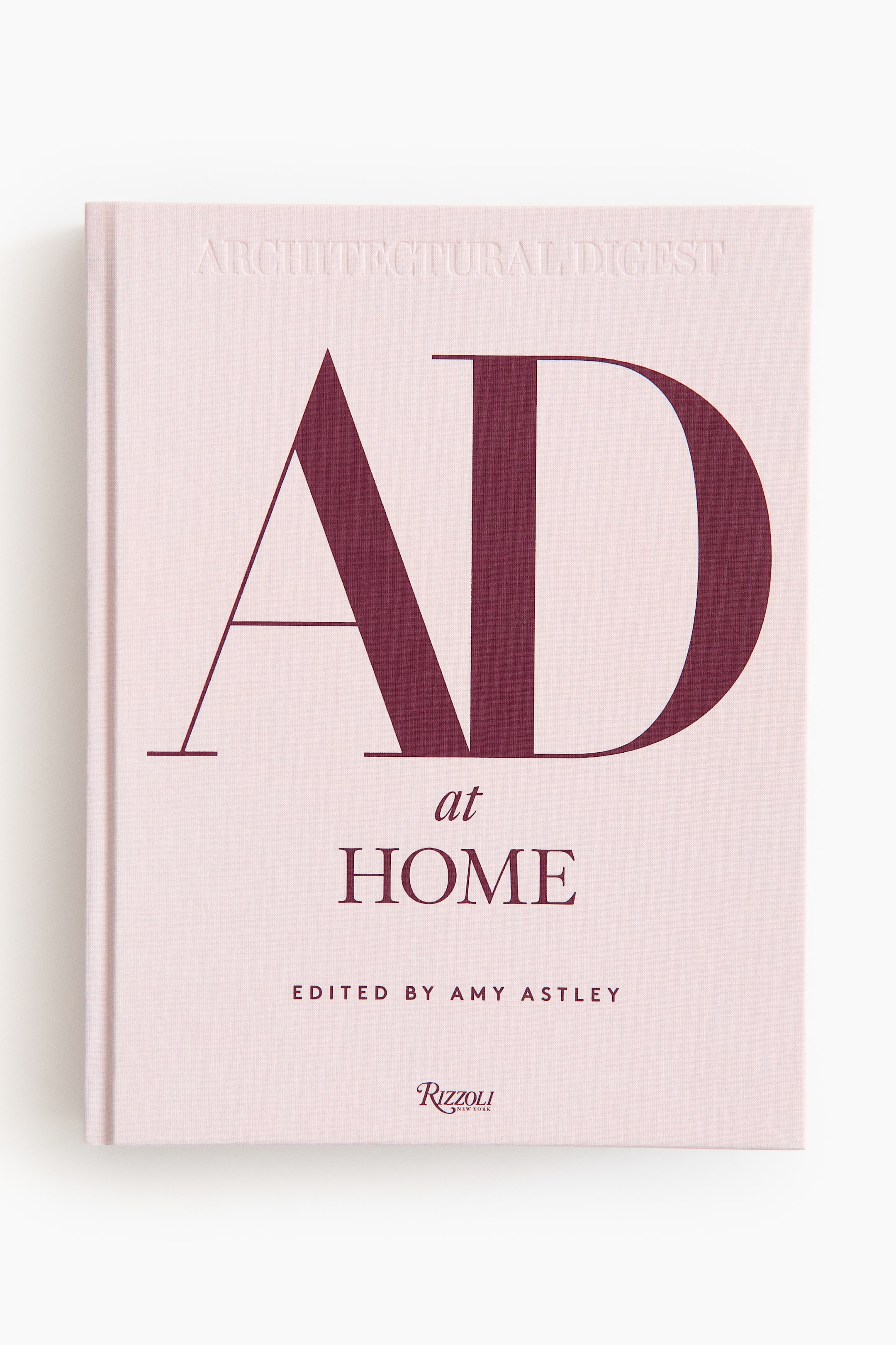 Visa större bild: "Ad At Home: Architectural Digest-boken står framåt och visar sitt ljusrosa tygbundna omslag. Omslaget pryds av stora, mörkröda 'AD' och 'at HOME'-texter, med 'REDAKTÖR AMY ASTLEY' och 'Rizzoli New York'-logotypen också i mörkrött, medan den präglade 'ARCHITECTURAL DIGEST'-texten framträder subtilt högst upp."