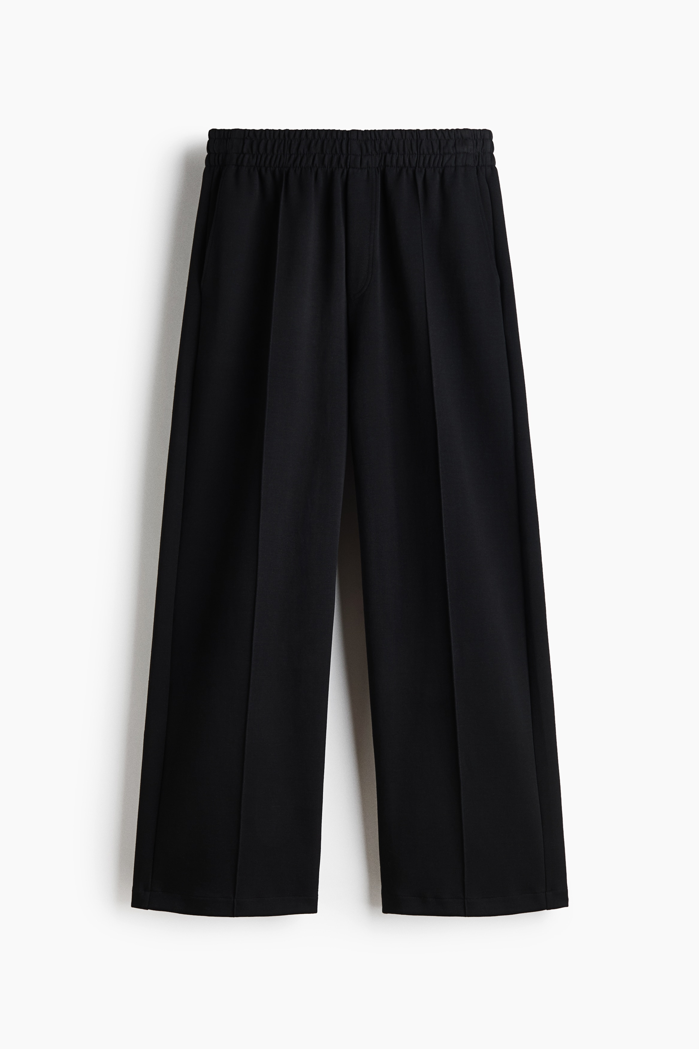 Dark brown track pants featuring a relaxed fit, straight legs and prominent sewn-in creases down the front are worn by the model. The model is standing with hands tucked into the diagonal side pockets, styling the trousers with a grey zip-up jacket, a light grey jumper draped over the shoulders, and black shoes.
