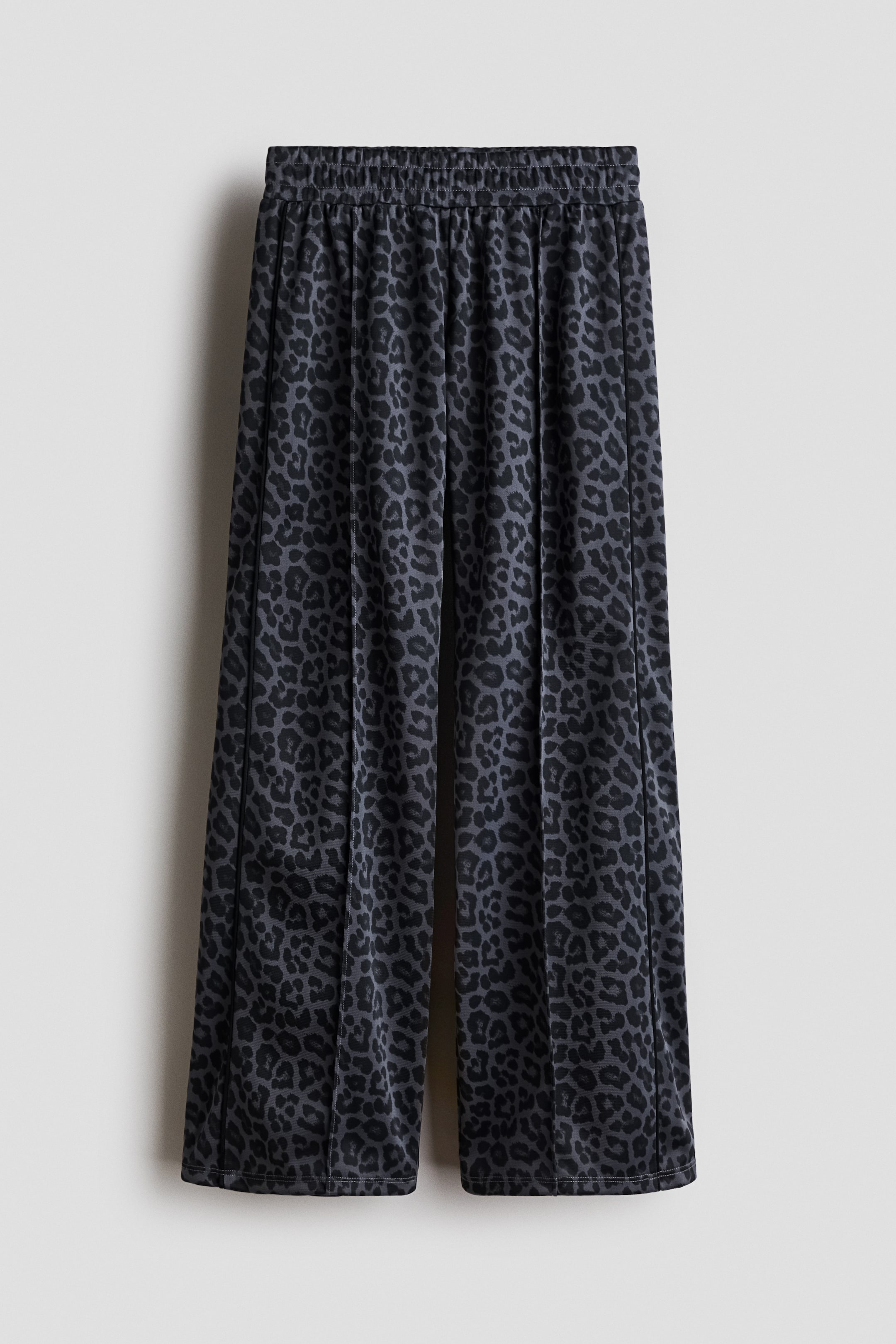 View larger image: FRONT track pants are displaying a dark grey and black leopard print with a slight sheen. They feature an elasticized waist, wide legs, and sewn-in creases down the front, which are highlighted by contrasting dark piping along the creases and outer sides.