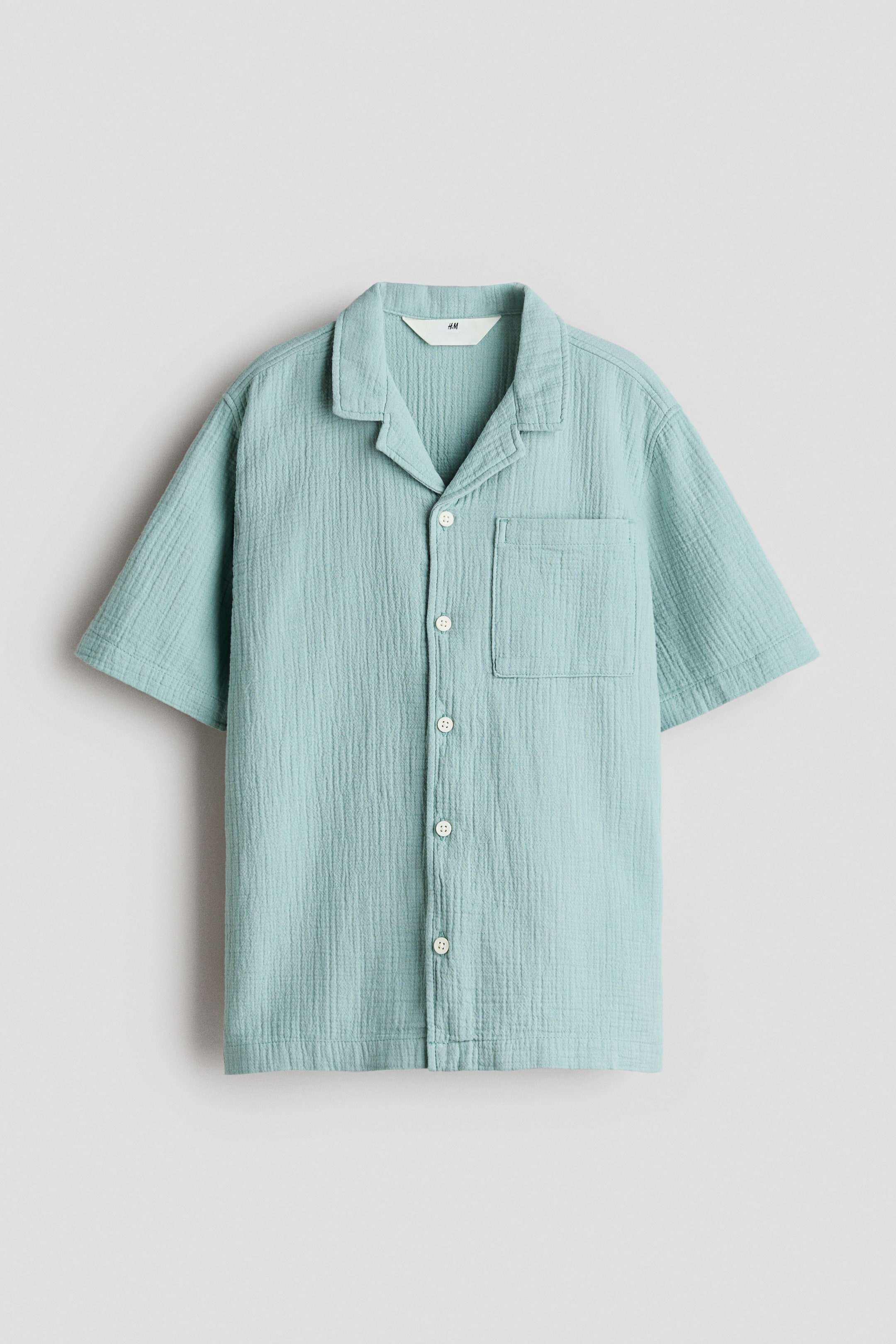 View larger image: A light blue-green, loose-fit, short-sleeved shirt is facing front, showcasing its soft cotton muslin texture. It features a resort collar, buttons down the front, a patch chest pocket, gently dropped shoulders, and a straight-cut hem.