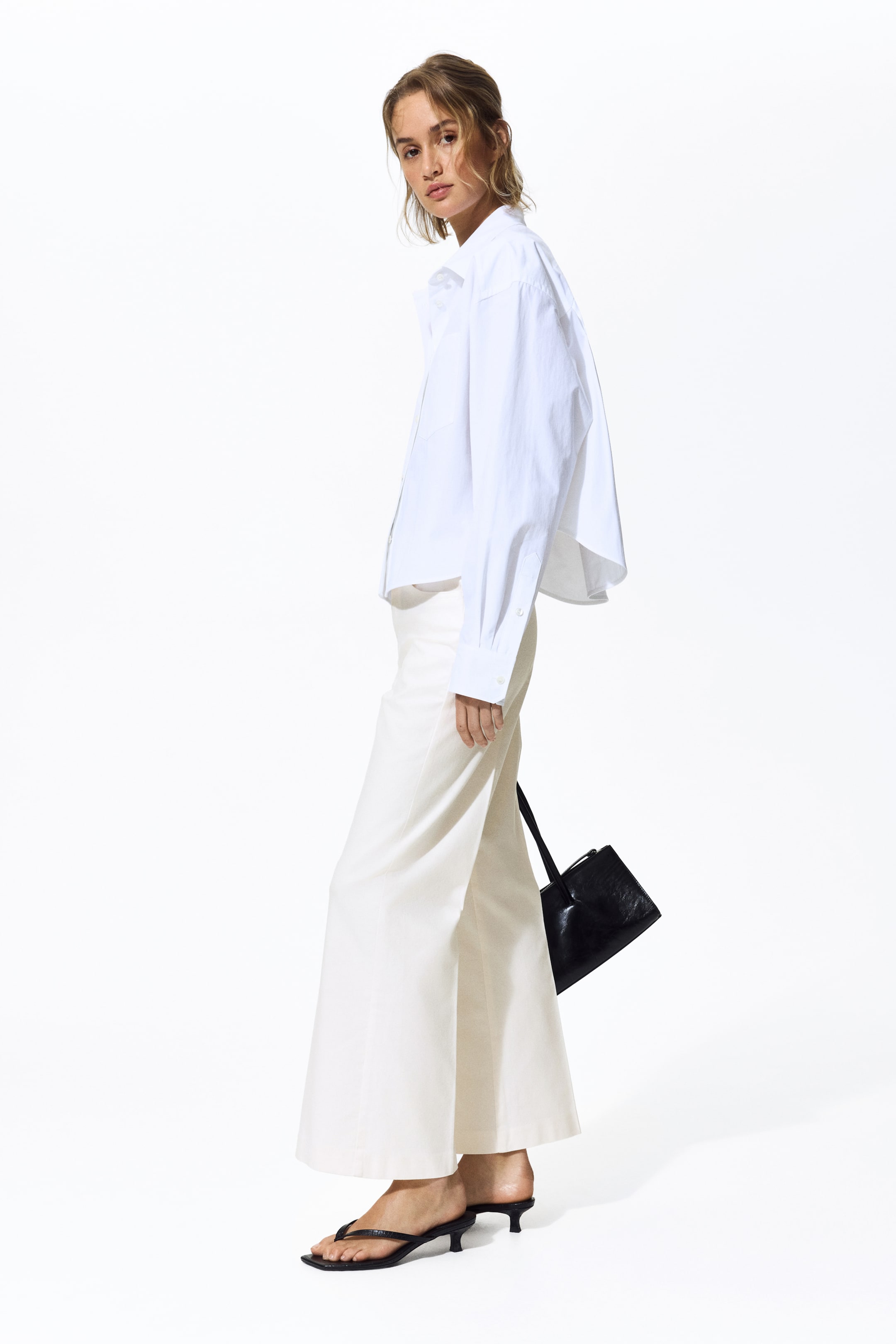 Преглед на по-голямо изображение: Бяла риза с големи размери и копчета е представена от страничен ъгъл, с яка, дълги ръкави с маншети с копчета и скъсен подгъв. Тя е съчетана с кремави панталони с широки крачоли, черни сандали с тънък ток и квадратен връх, и черна структурирана чанта, която виси на презрамката си.