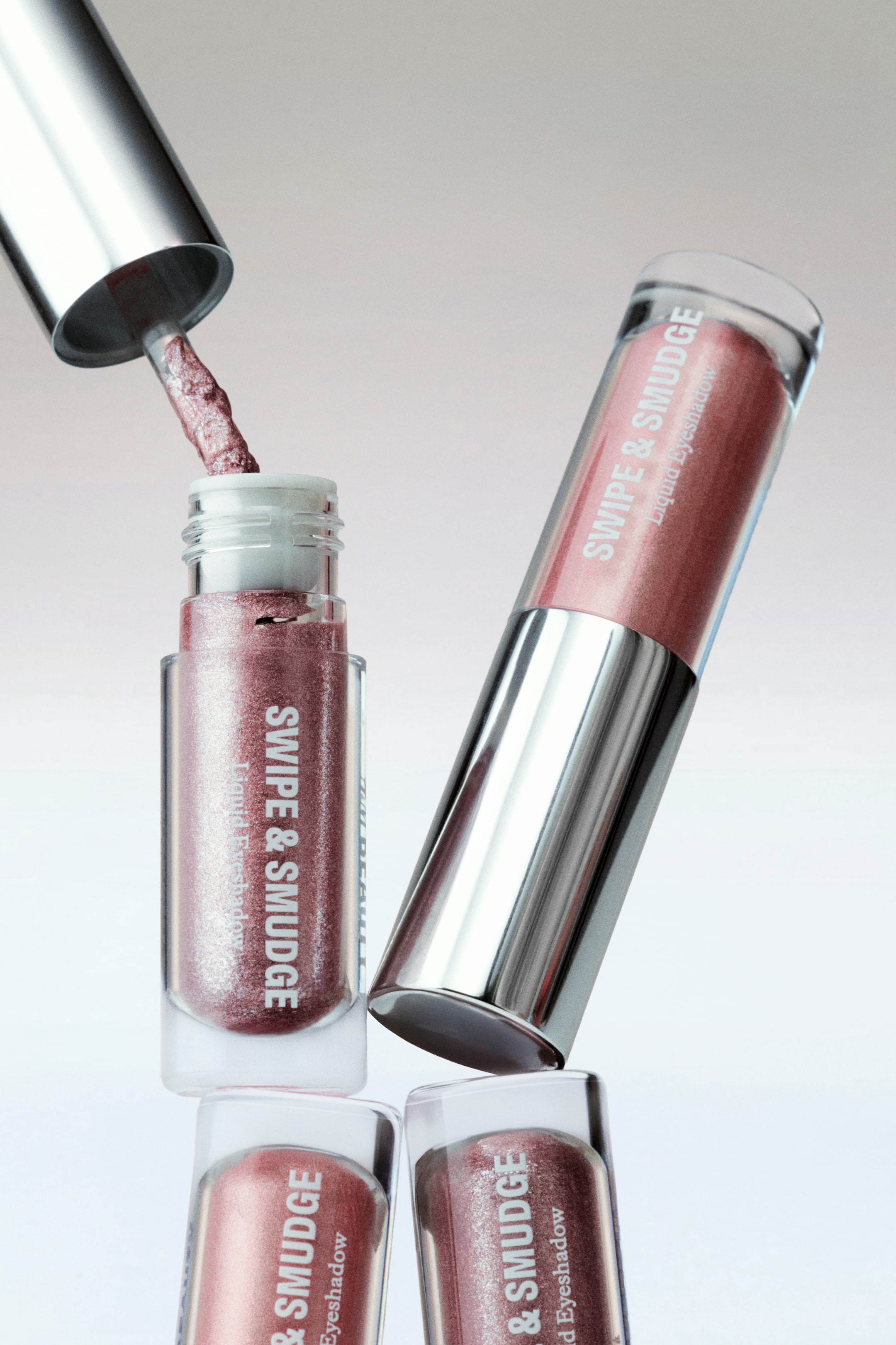 Visualizza immagine più grande: "Swipe & Smudge Liquid Eyeshadow viene presentato con un tubo trasparente aperto, il suo applicatore argentato che emerge e lascia gocciolare il prodotto rosato-marrone scintillante di nuovo nel tubo. Un tubo identico chiuso con un tappo argentato è appoggiato contro quello aperto, inclinato verso destra, mentre altri due tubi chiusi sono impilati sotto."