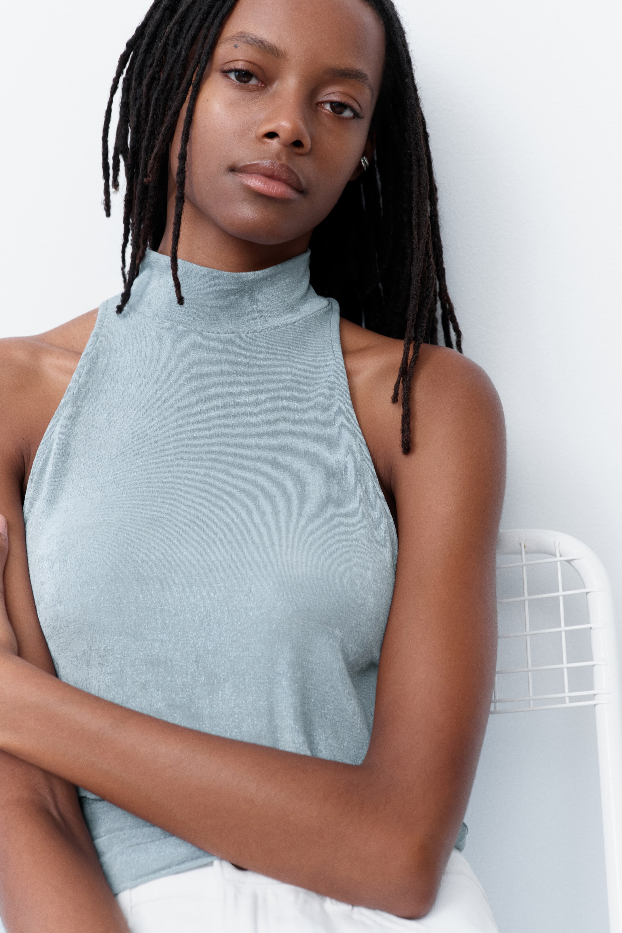 Ampliar la imagen: La modelo lleva una blusa de jersey azul claro, sin mangas, con cuello alto y recto, y un corte estrecho en los hombros. La blusa está estilizada por dentro de un pantalón blanco.