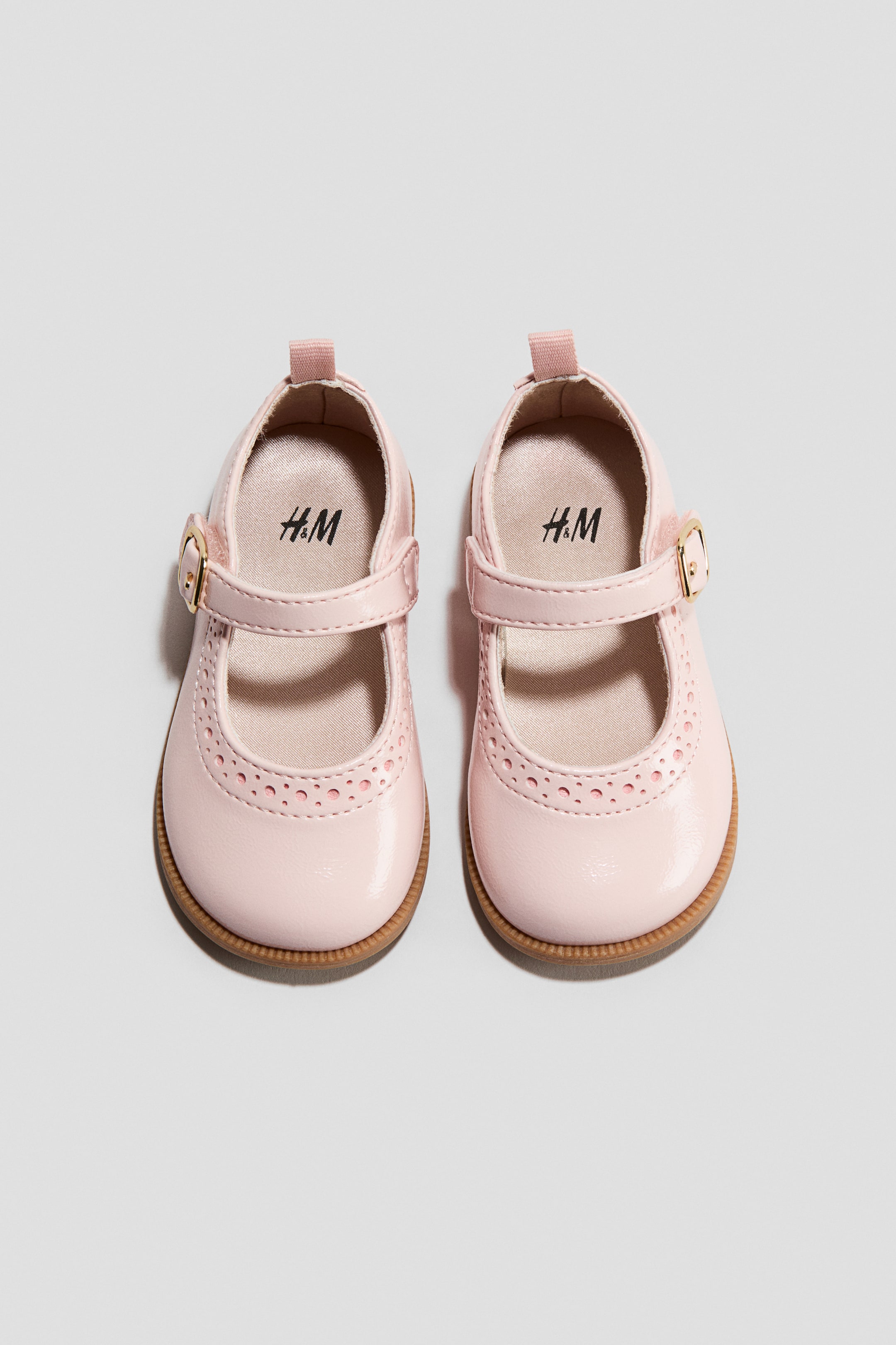 Ampliar la imagen: Un par de Mary Janes rosadas, presentado al FRENTE, presenta un tejido brillante recubierto con un borde perforado en la parte frontal. Cada zapato muestra una correa para el pie con una hebilla de color dorado, un forro de satén suave y un logo de H&M visible en la plantilla.