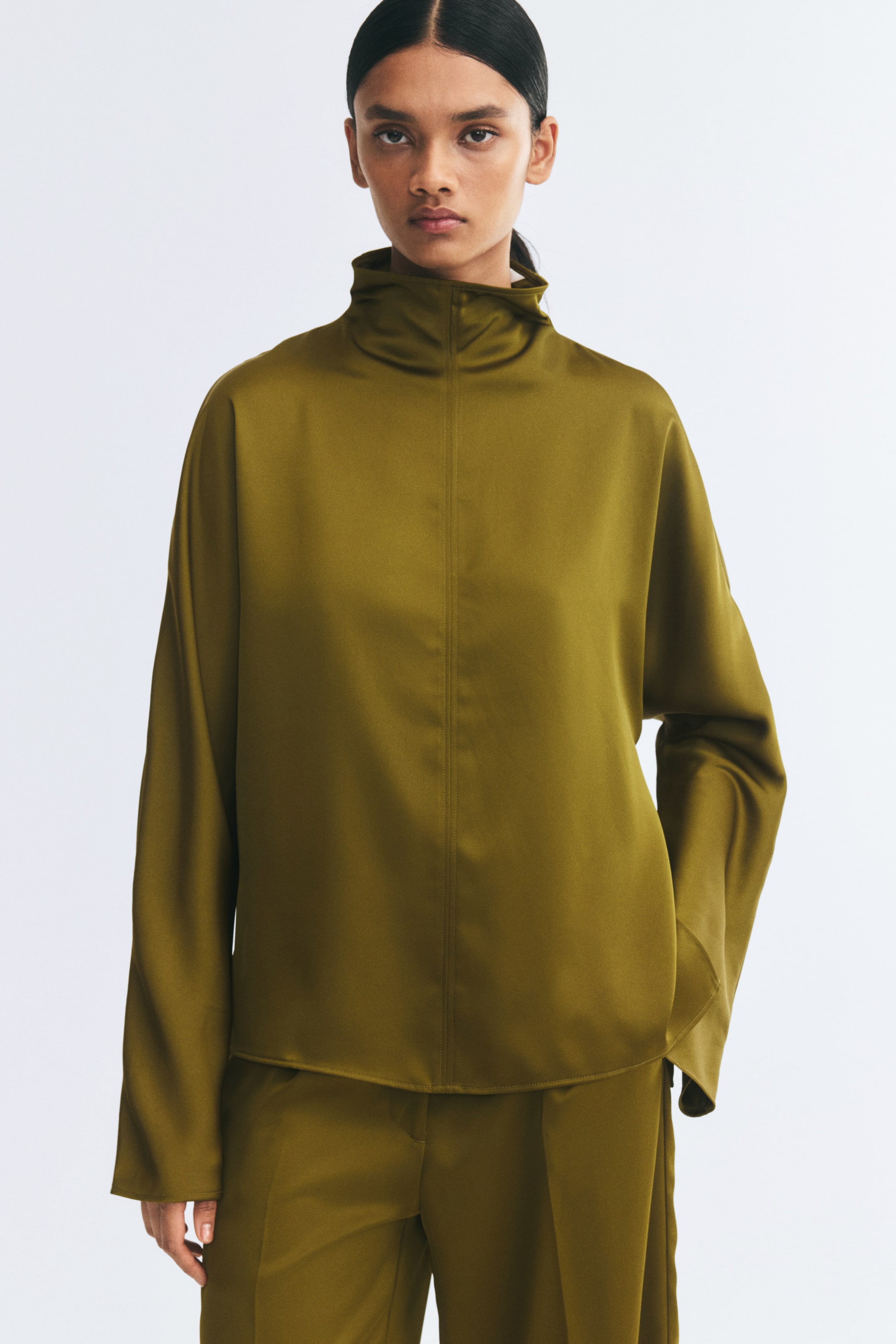 Agrandir l'image: Le mannequin porte un chemisier en satin vert olive à manches longues et col montant, avec une couture verticale visible au centre sur le devant, assorti à un pantalon coordonné.