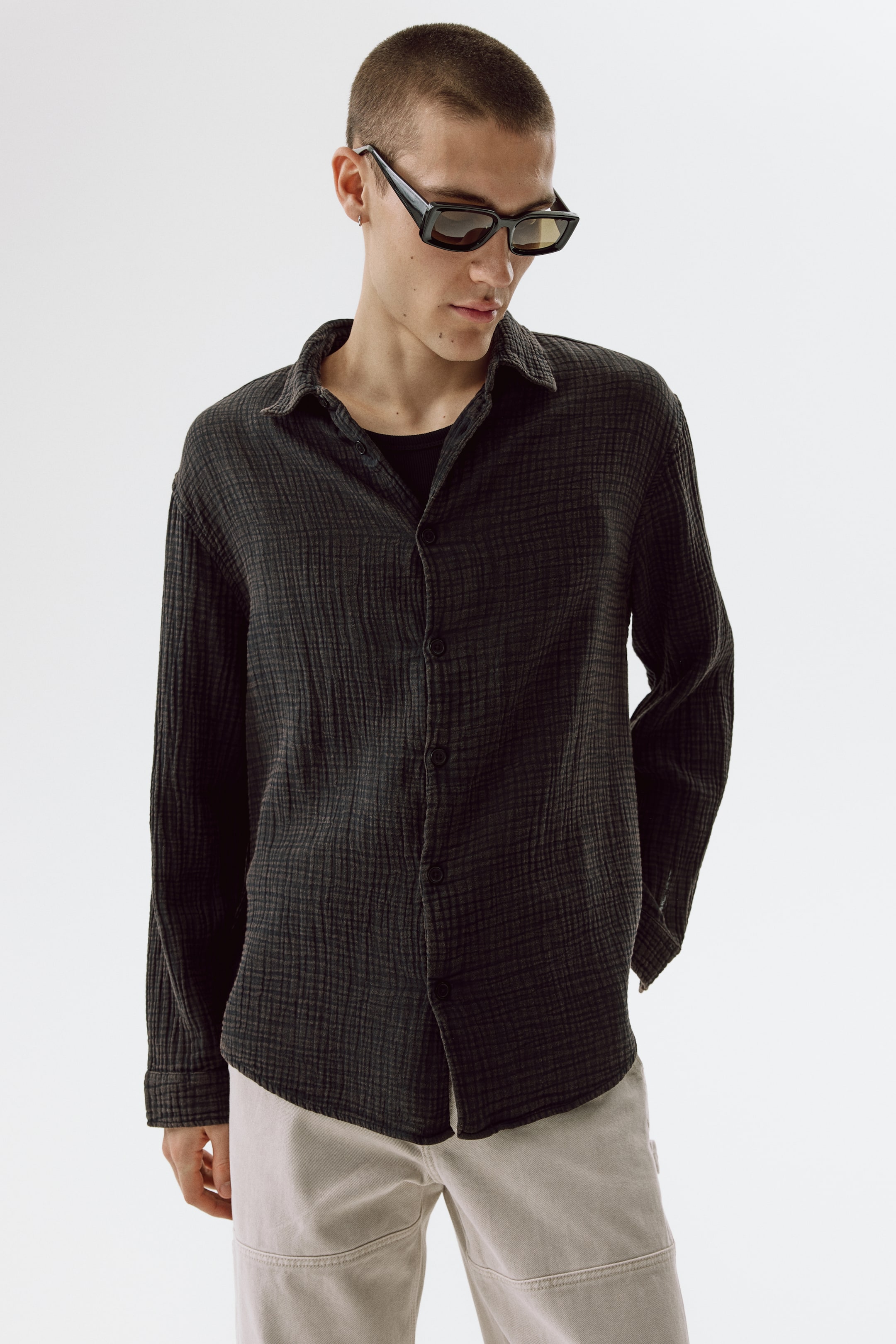 View larger image: The model is sporting a dark grey cotton muslin shirt with a washed appearance, layered over a black tee. This shirt features a collar, visible front buttons, long sleeves with buttoned cuffs, and a subtly rounded hem, with some front buttons left undone.