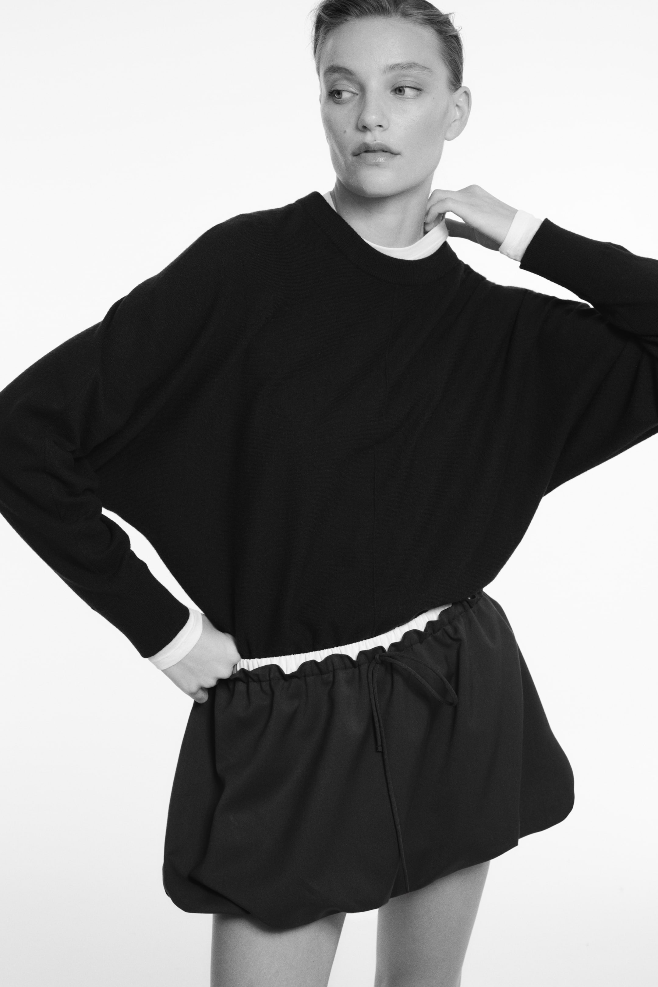 Agrandir l'image: Ce pull en maille noir est présenté de face, avec une coupe décontractée, un col rond bordé de côtes et une couture verticale visible courant au centre. Les manches longues se terminent par des poignets côtelés, laissant entrevoir un col et des poignets blancs provenant d’un sous-vêtement superposé.
