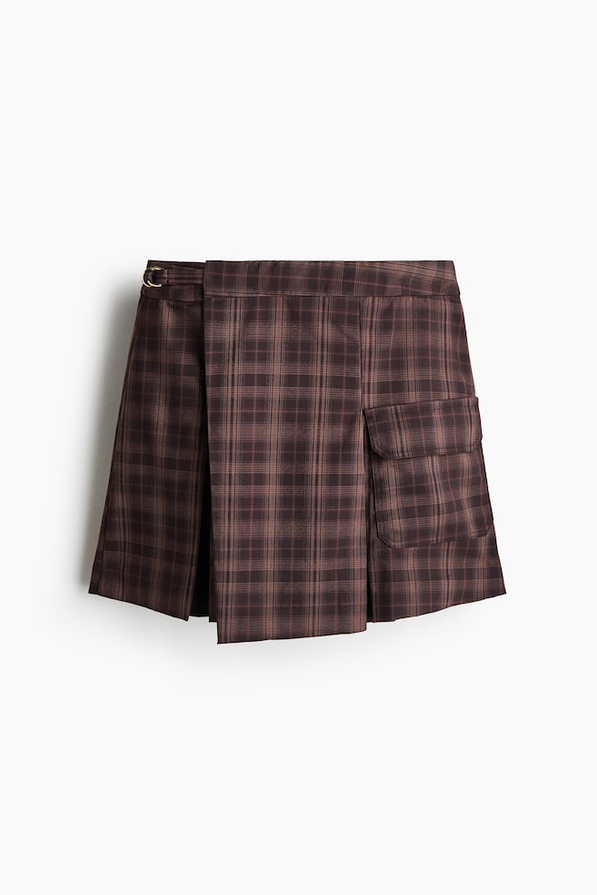 A brown plaid wrap skirt is shown from the front, featuring an asymmetrical wrap-around panel with a small metal buckle fastening at the side. The skirt also features a cargo-style flap pocket on the left side and structured pleating. - 1