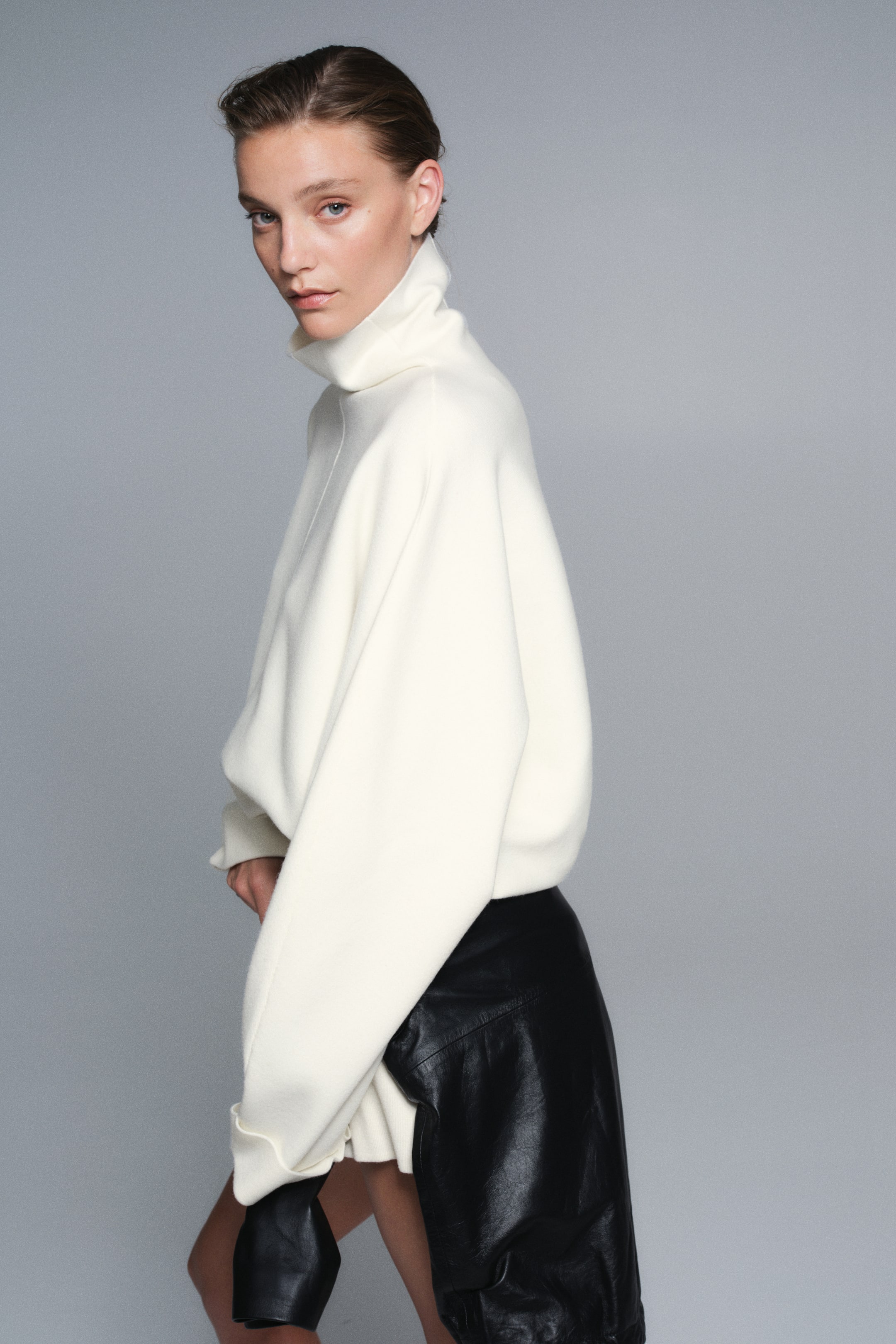 Visualizza l'immagine ingrandita: Un maglione oversize in maglia color crema è indossato rivolto a sinistra, mettendo in risalto il collo alto, una prominente cucitura centrale sul davanti e ampie maniche a pipistrello con polsini ripiegati. Il maglione cade morbido sopra un capo in pelle nera, evidenziandone la texture soffice a maglia fine e la silhouette rilassata.