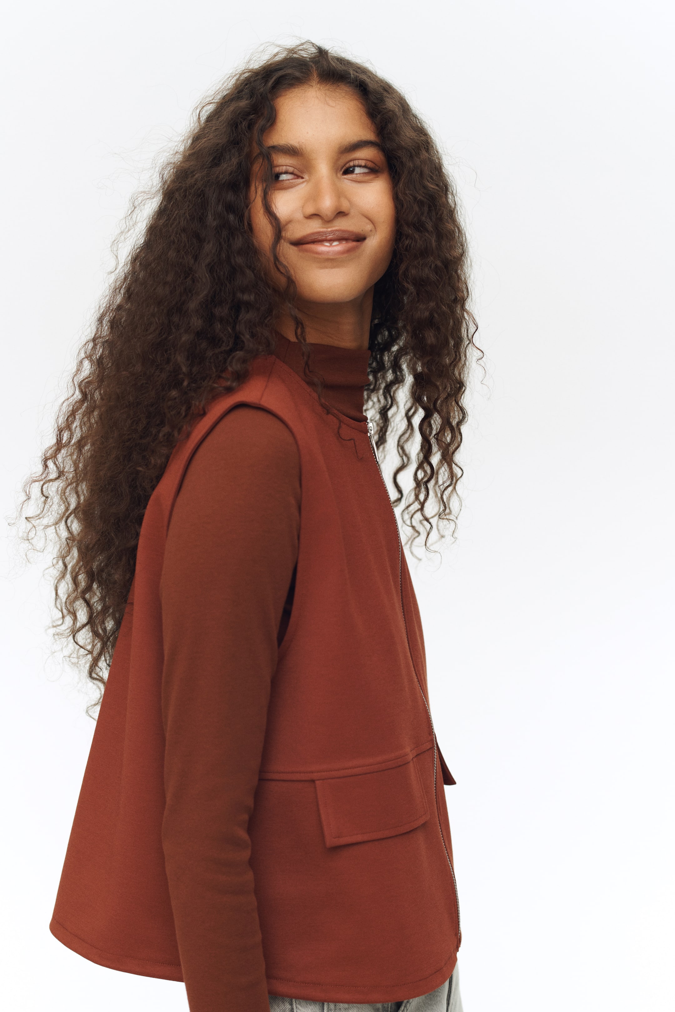 Ampliar la imagen: Un chaleco sin mangas de color óxido se superpone sobre un top de cuello alto de manga larga a juego, ambos mostrados desde un ángulo de tres cuartos. El chaleco está diseñado con un cierre de cremallera plateada y un bolsillo con solapa rectangular en su parte frontal.