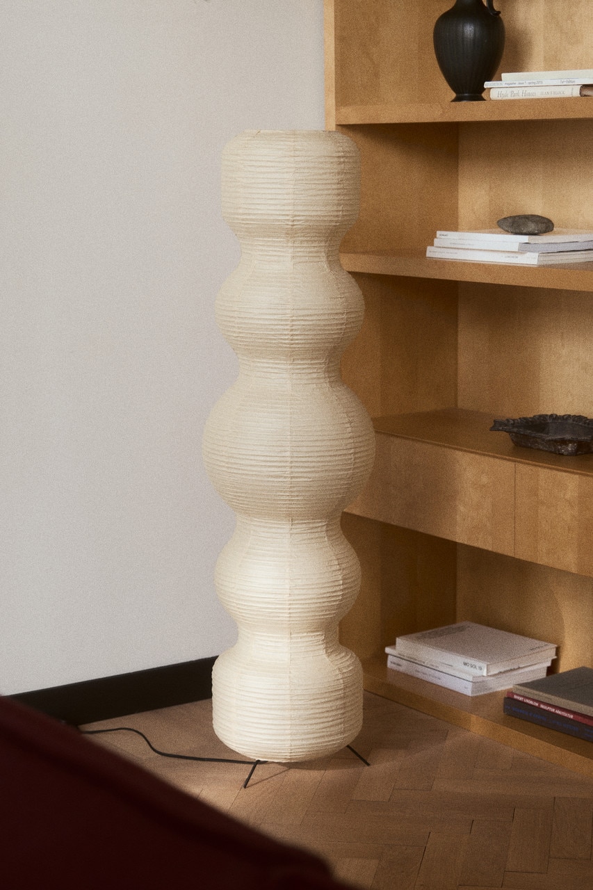 A floor lamp is standing FRONT, featuring a cream-colored rice paper shade with a stacked, organic, multi-lobed shape and horizontal pleats. A black three-legged metal base is supporting the shade, and a black electrical cord is extending from its bottom left.