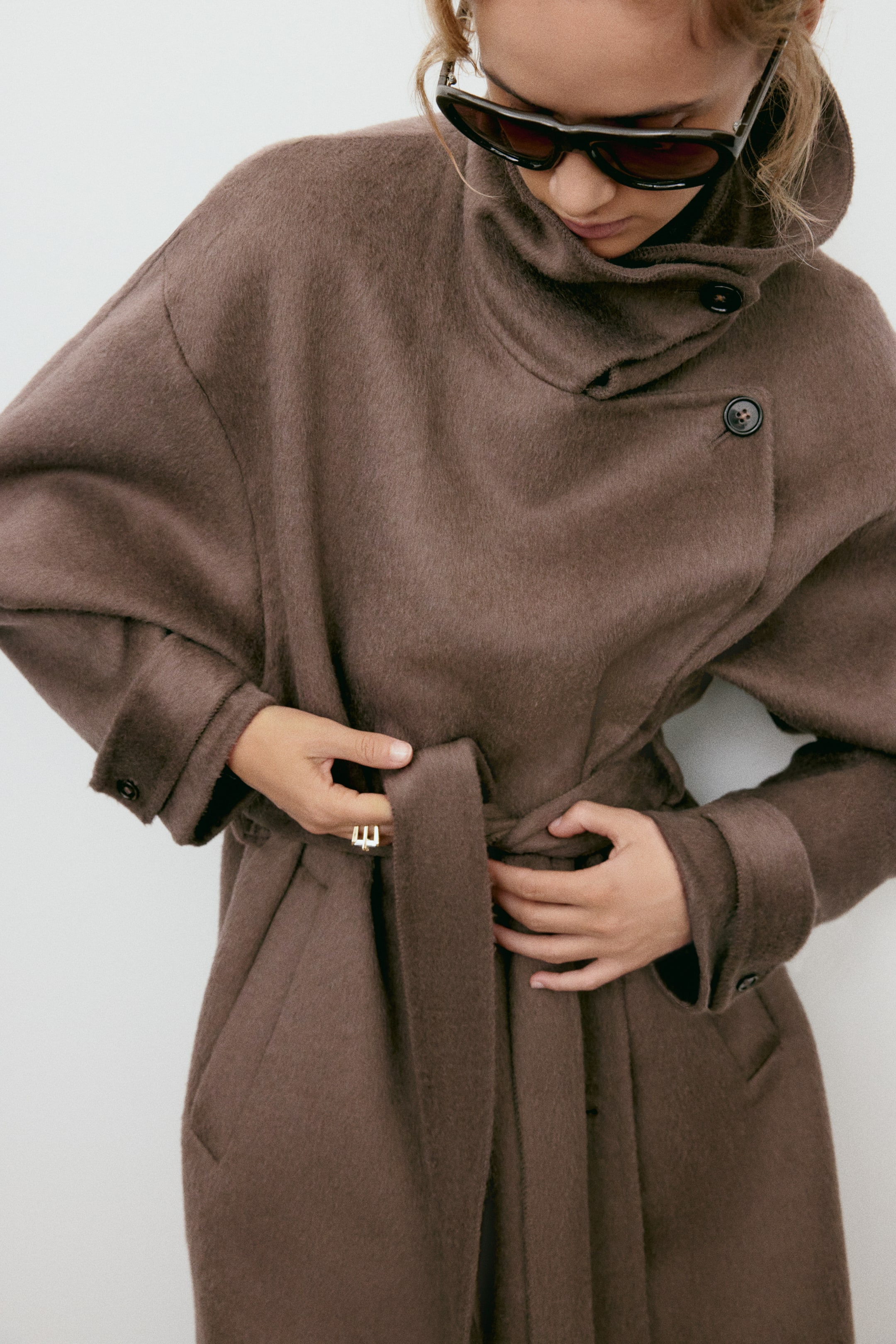 Zobraziť väčší obrázok: Modelka má na sebe hnedý oversized kabát z plstenej vlnenej zmesi a upravuje si zodpovedajúci viazací opasok v páse. Kabát má vysoký lievikovitý golier s gombíkovým zapínaním, spustené ramenné švy a nastaviteľné gombíkové pásiky na manžetách.