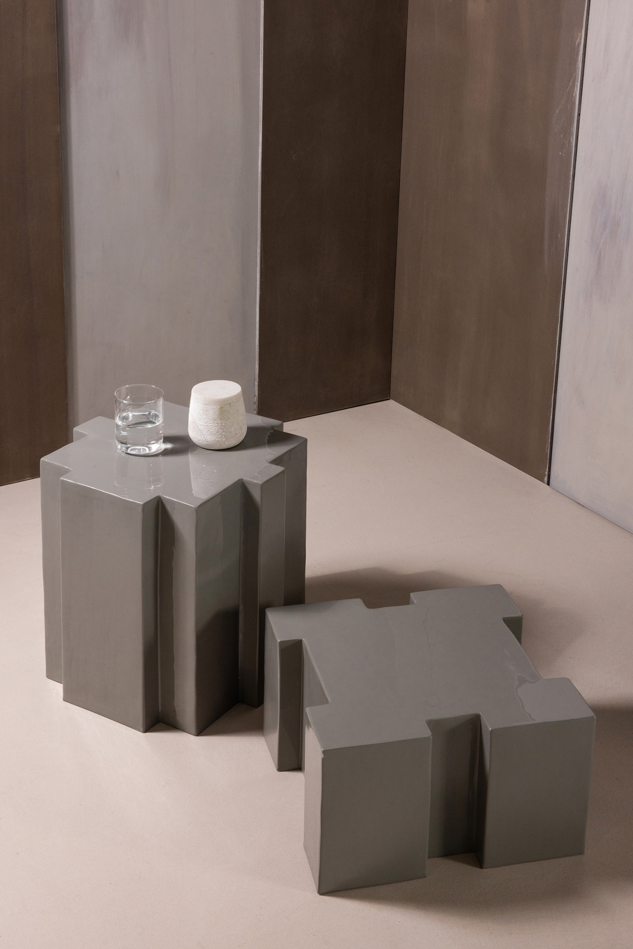 Agrandir l'image: Deux tables géométriques grises sont présentées. La table la plus haute, dotée d'une finition brillante et d'un plateau octogonal avec des angles rentrants, est debout, sa base formée de panneaux verticaux, et supporte un verre d'eau clair et un petit objet texturé blanc cassé. La table plus basse, également avec une finition brillante et un plateau en forme de H avec des pieds en bloc, repose sur une surface plane à côté.