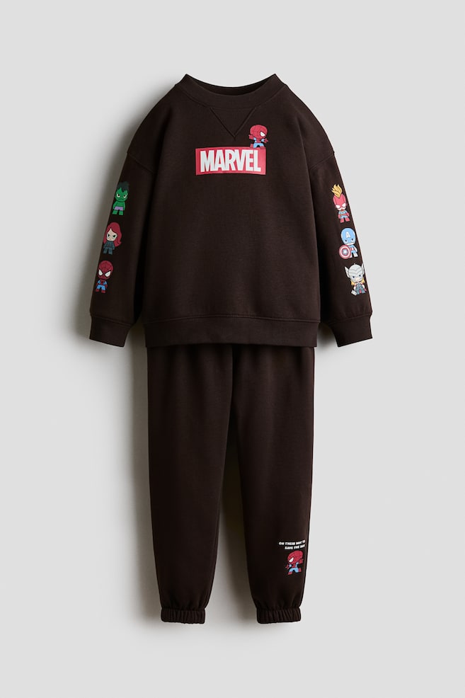 Um conjunto de fato de treino da Marvel em castanho escuro está a apresentar uma vista frontal. A sweatshirt possui uma gola redonda canelada, punhos canelados e uma bainha canelada, exibindo proeminentemente um logótipo 'MARVEL' em vermelho e branco na frente, com uma pequena personagem do Homem-Aranha a espreitar por cima, e várias pequenas ilustrações de personagens da Marvel a adornar ambas as mangas. As calças de fato de treino a condizer apresentam punhos elásticos, com uma pequena personagem do Homem-Aranha e texto acompanhante visíveis na parte inferior da perna esquerda." - 1