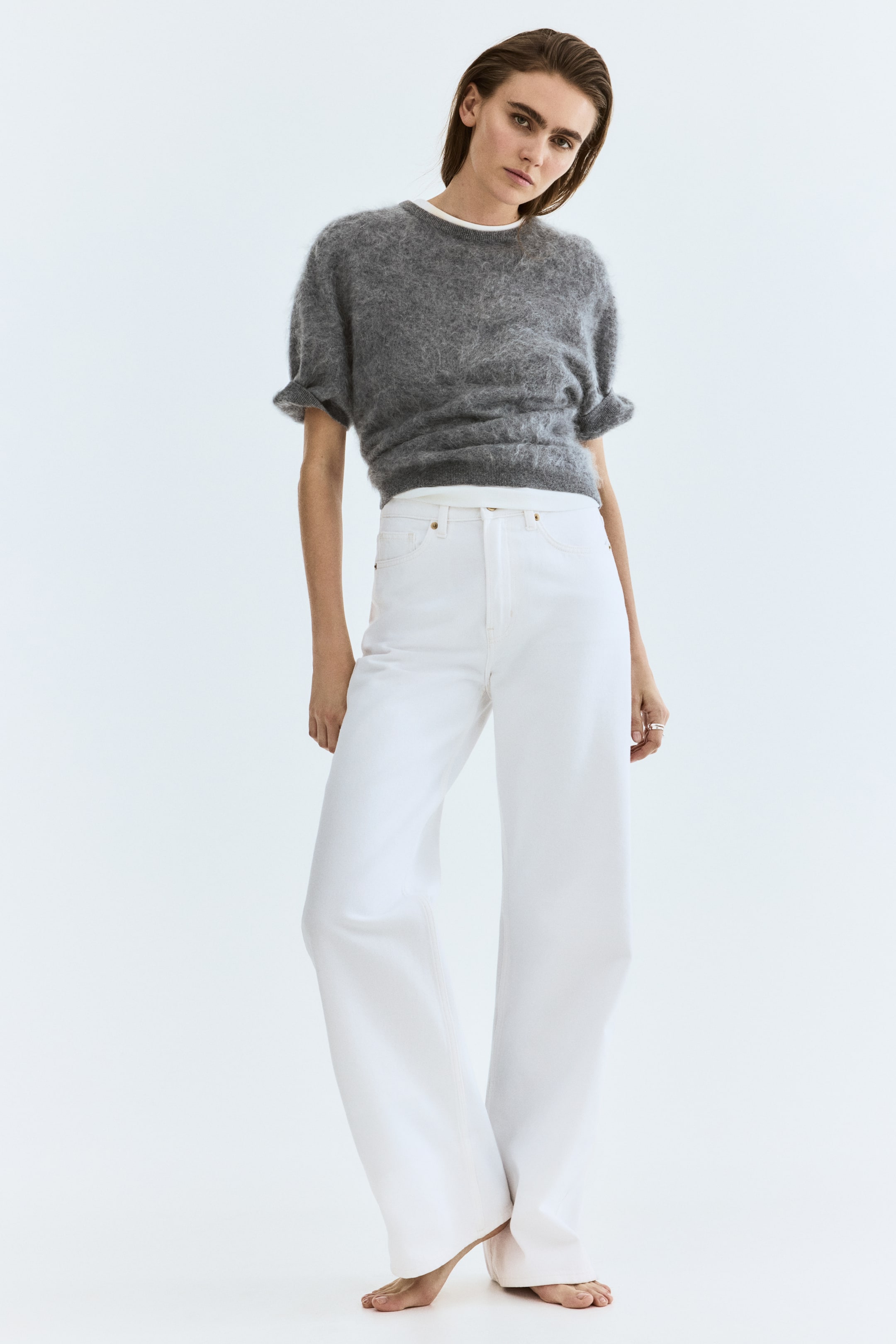 Xem ảnh lớn hơn: Một chiếc áo len xám mờ ngắn tay, có cổ tròn, tay áo xếp ly và chiều dài lửng, được mặc bên ngoài một chiếc áo trắng. Quần jean trắng cạp cao ống rộng, hiển thị túi trước rõ ràng và đường may nhẹ, được mặc bên dưới áo len.
