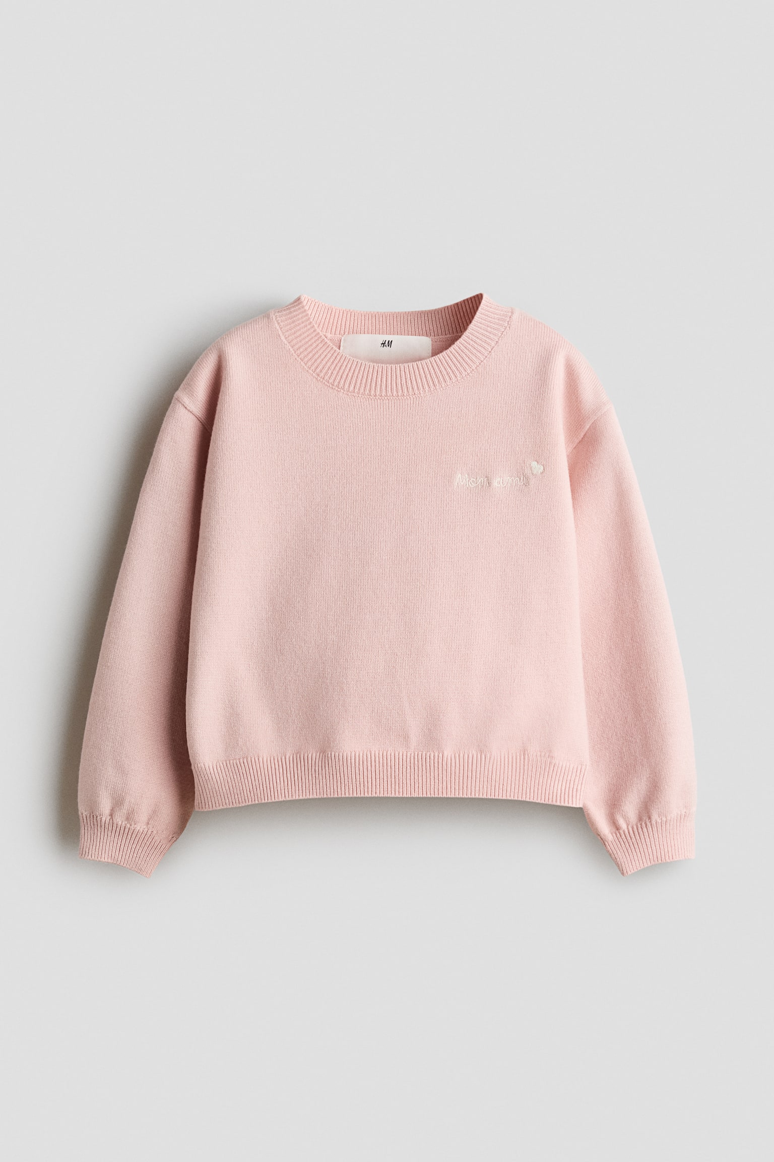 A light pink, fine-knit jumper is shown from the front, displaying a round, rib-trimmed neckline, dropped shoulders, and ribbed cuffs and hem. The jumper features small white embroidered text and a heart detail on the left chest.