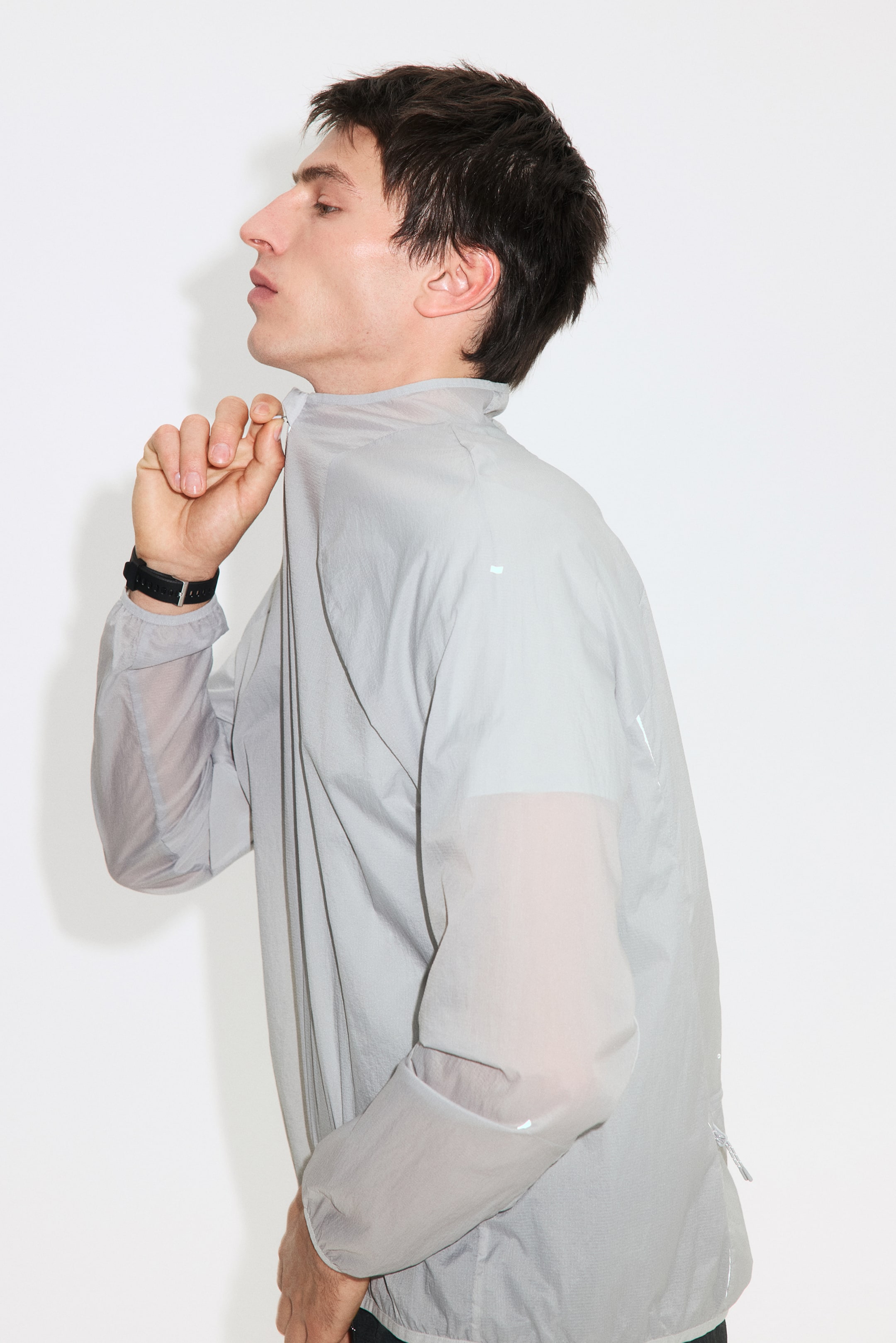 Agrandir l'image: Une veste gris clair semi-transparente est présentée de profil, tournée vers la gauche, avec une main tirant sa fermeture éclair avant, formant un col montant. Des manches longues s'étendent de la veste, et de petits éléments réfléchissants apparaissent sur son corps et ses manches, avec un ourlet de manche élastiqué et une poche zippée également visible sur son côté inférieur.