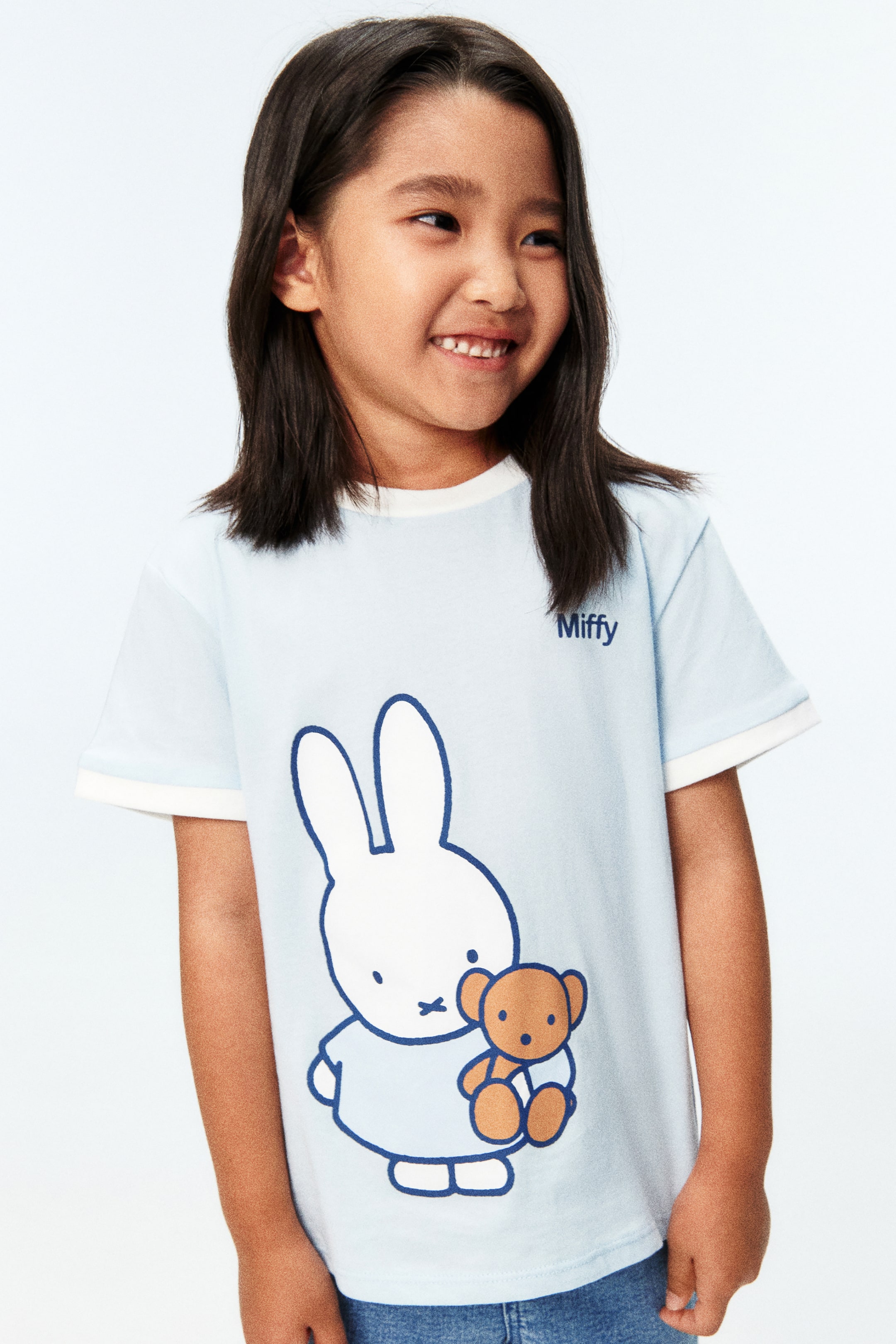 View larger image: A light blue T-shirt, featuring a front print of a white rabbit holding a brown teddy bear and "Miffy" text, is worn by the model. It has a round neckline with contrasting white trims at the neckline and cuffs.
