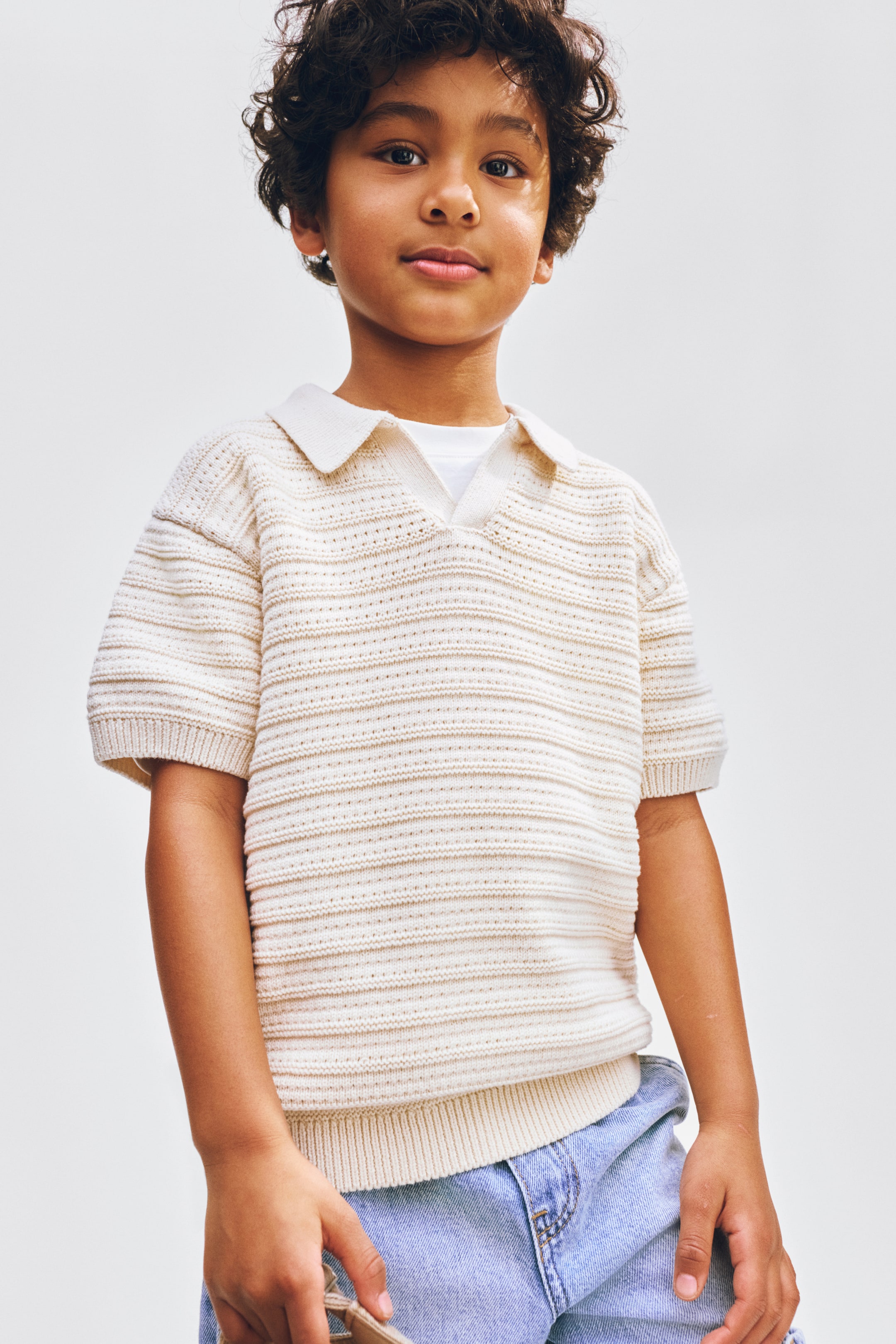 Grotere afbeelding bekijken: Een crèmekleurige gebreide poloshirt met een klassieke kraag en een V-hals opening, afwisselende horizontale gestructureerde strepen met effen en geperforeerde delen, korte mouwen met geribde boorden en een geribde zoom, gedragen over een wit onderhemd.