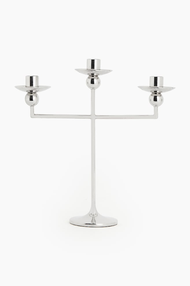 Een metalen kandelaar met drie armen staat rechtop en toont van voren zijn slanke silhouet en gepolijste zilveren afwerking. Het ontwerp heeft een slanke centrale steel die uit een uitlopende ronde voet oprijst en drie kaarshouders draagt, elk voorzien van een bolvormig detail onder een ondiepe druipschaal. - 1