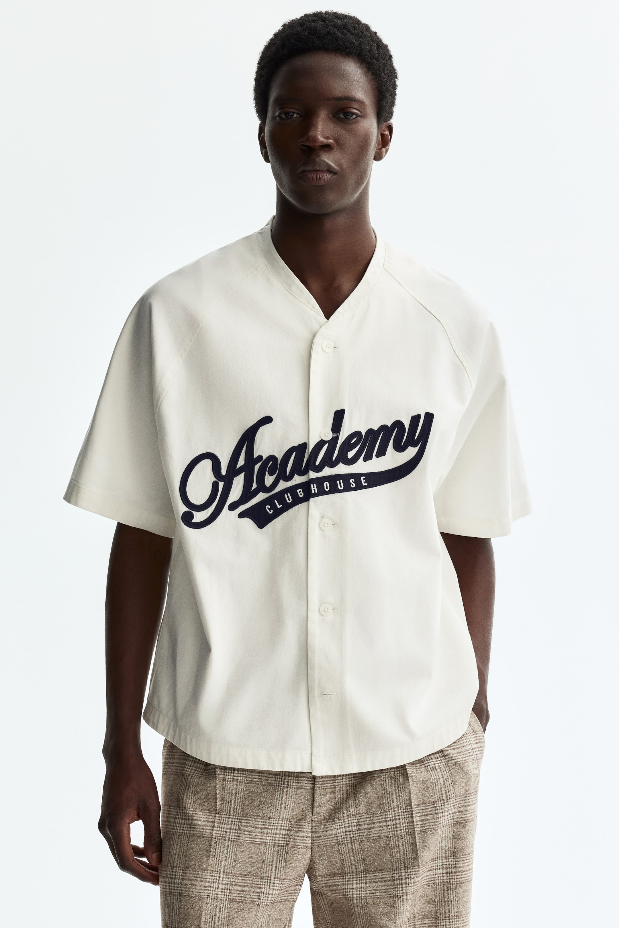 View larger image: An off-white baseball shirt is shown from the front, featuring short raglan sleeves and a collarless V-neck. A dark blue 'Academy Clubhouse' graphic is appliquéd on the front, and it fastens with white buttons down the center.