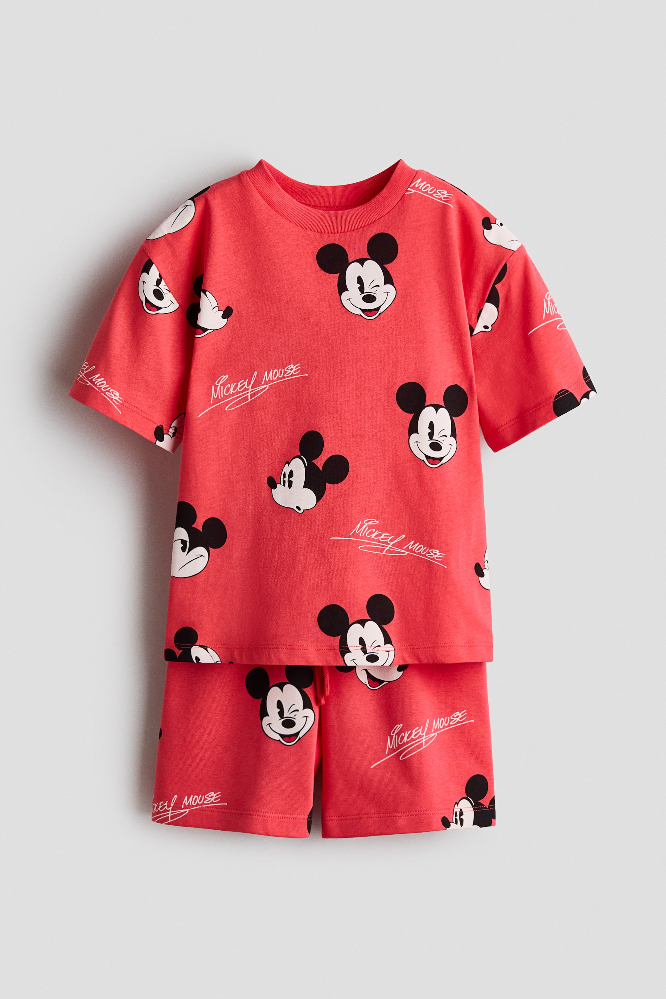 Größeres Bild ansehen: Ein passendes rotes T-Shirt und Shorts-Set zeigt seine Vorderansicht, bedeckt mit einem Allover-Print von Mickey Mouse-Gesichtern und weißen "Mickey Mouse"-Signaturen. Das T-Shirt verfügt über einen gerippten Rundhalsausschnitt, weite kurze Ärmel und überschnittene Schultern, und die Shorts zeigen eine sichtbare Kordel und eine verdeckte Elastik am Bund.