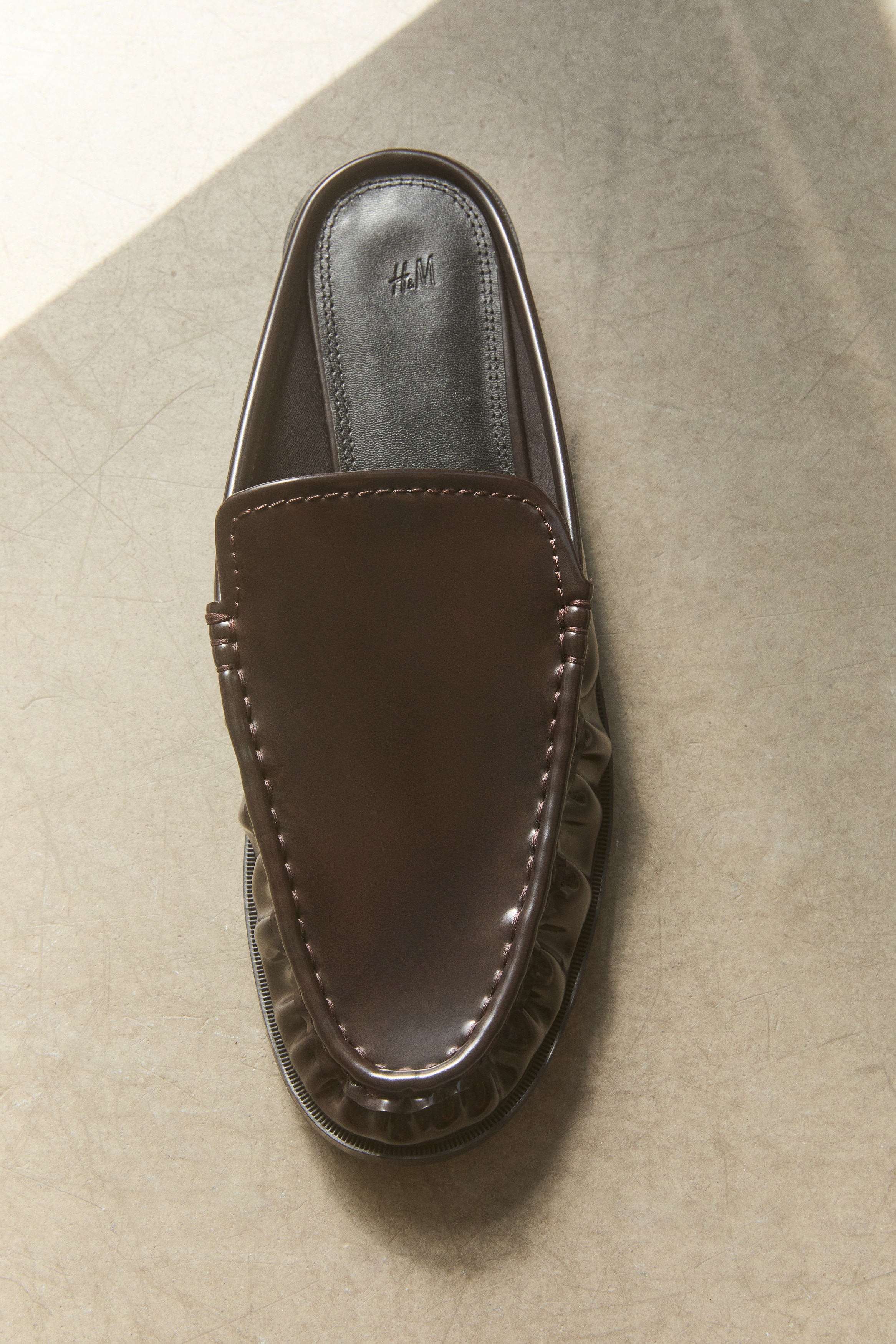 A pair of burgundy patent-finish backless loafers is showcased from a top-down perspective. They feature moccasin-style stitching and gathered detailing around the vamp.