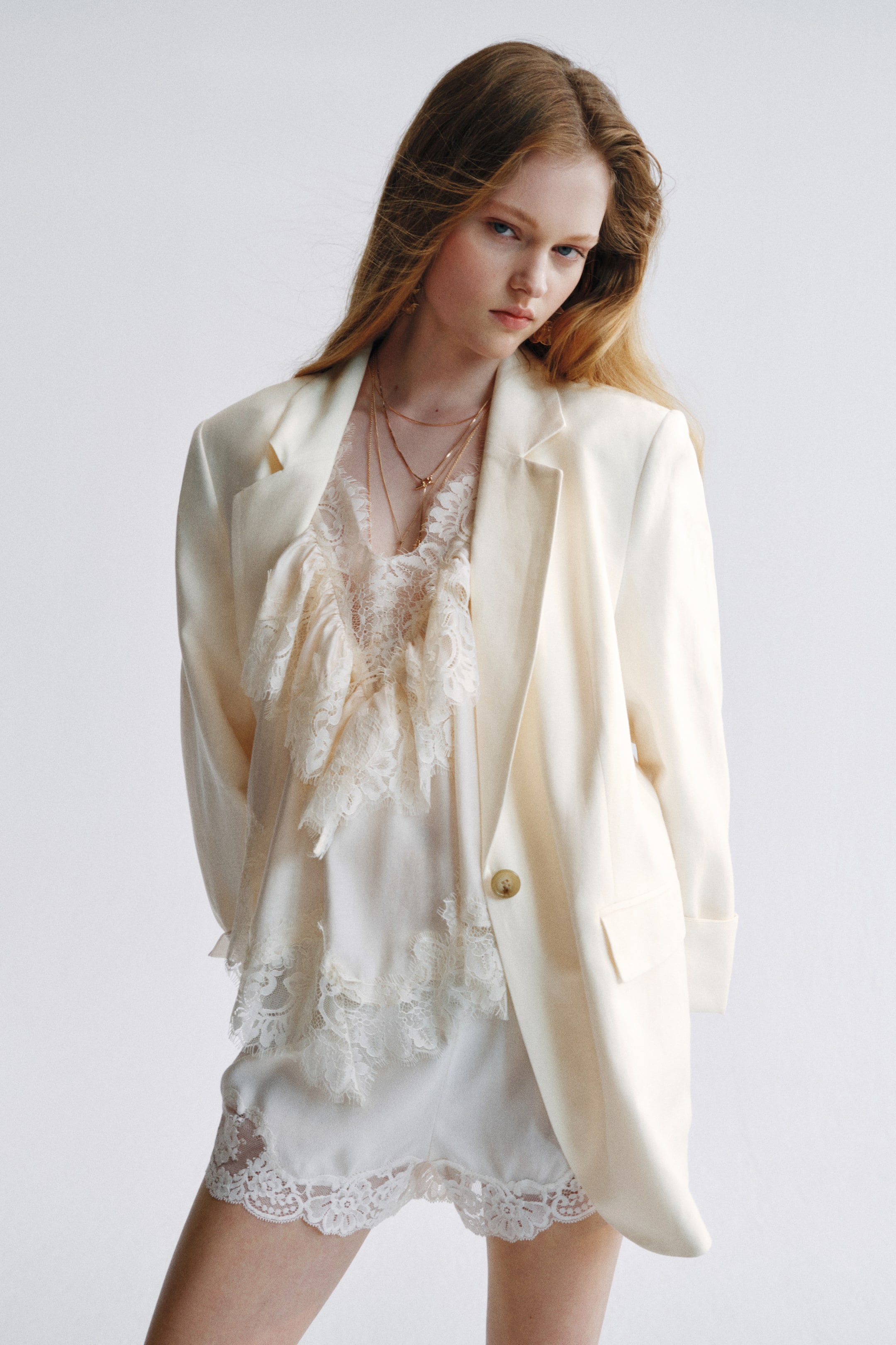 Ampliar la imagen: La modelo lleva un blazer crema de un solo botón con solapas de muesca y hombreras. Presenta un botón frontal visible y bolsillos con solapa en jet, superpuesto sobre un top de encaje crema, shorts a juego y múltiples collares dorados.
