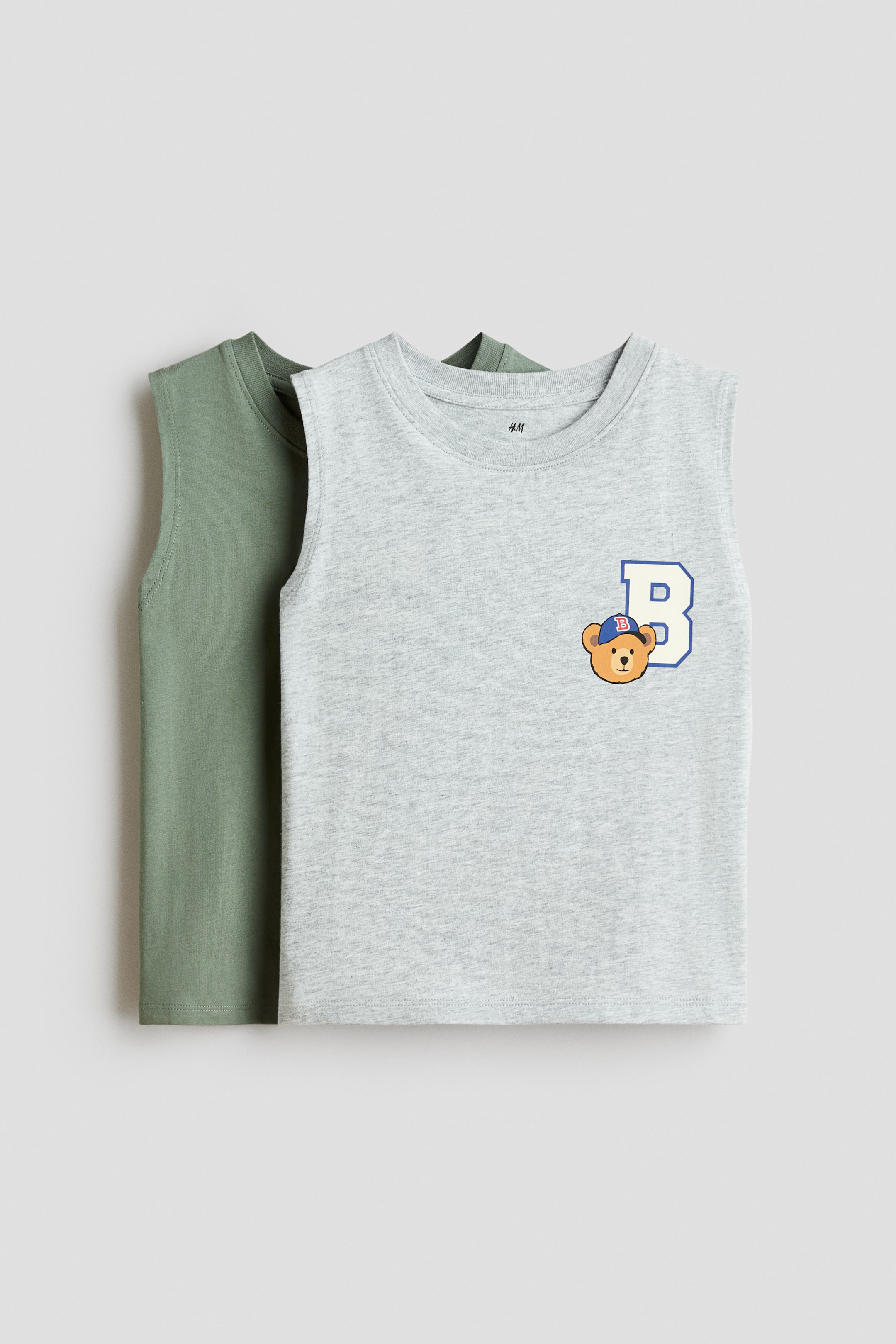 View larger image: Two cotton jersey tank tops are shown from the front, with a solid sage green tank layered behind a light grey melange tank. The grey tank features a round, rib-trimmed neckline and a graphic print on the left chest depicting a cartoon bear wearing a blue cap next to a blue letter "B."