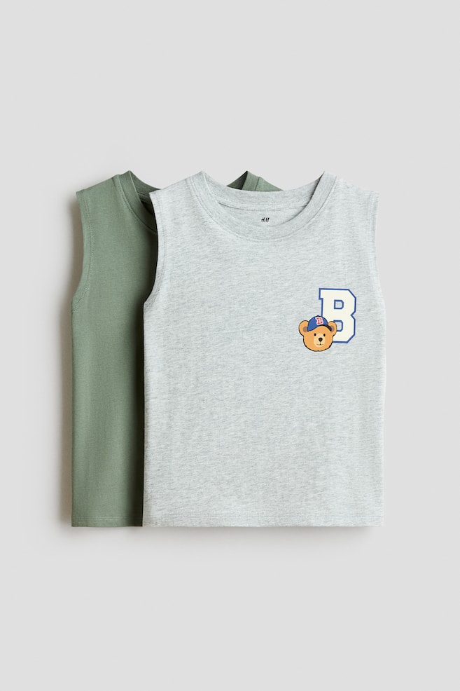 Two cotton jersey tank tops are facing the front, with a solid sage green tank layered behind a light grey melange tank. The grey tank is featuring a round, rib-trimmed neckline and a graphic print on the left chest depicting a cartoon bear wearing a blue cap next to a blue letter "B." - 1