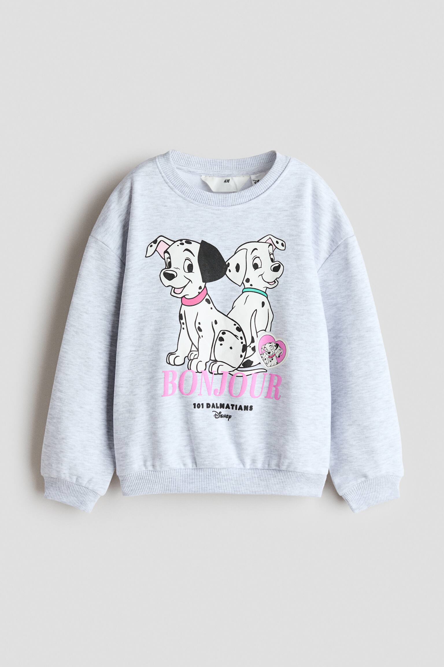 Ein hellgrau meliertes Sweatshirt ist von vorne zu sehen, mit Rundhalsausschnitt, Ã¼berschnittenen Schultern und langen Ãrmeln mit RippbÃ¼ndchen. Auf der Vorderseite ist ein gedrucktes Motiv von zwei Cartoon-Dalmatinern Ã¼ber dem Wort âBONJOURâ in pinker Schrift zu sehen, abgerundet durch einen Rippbund.