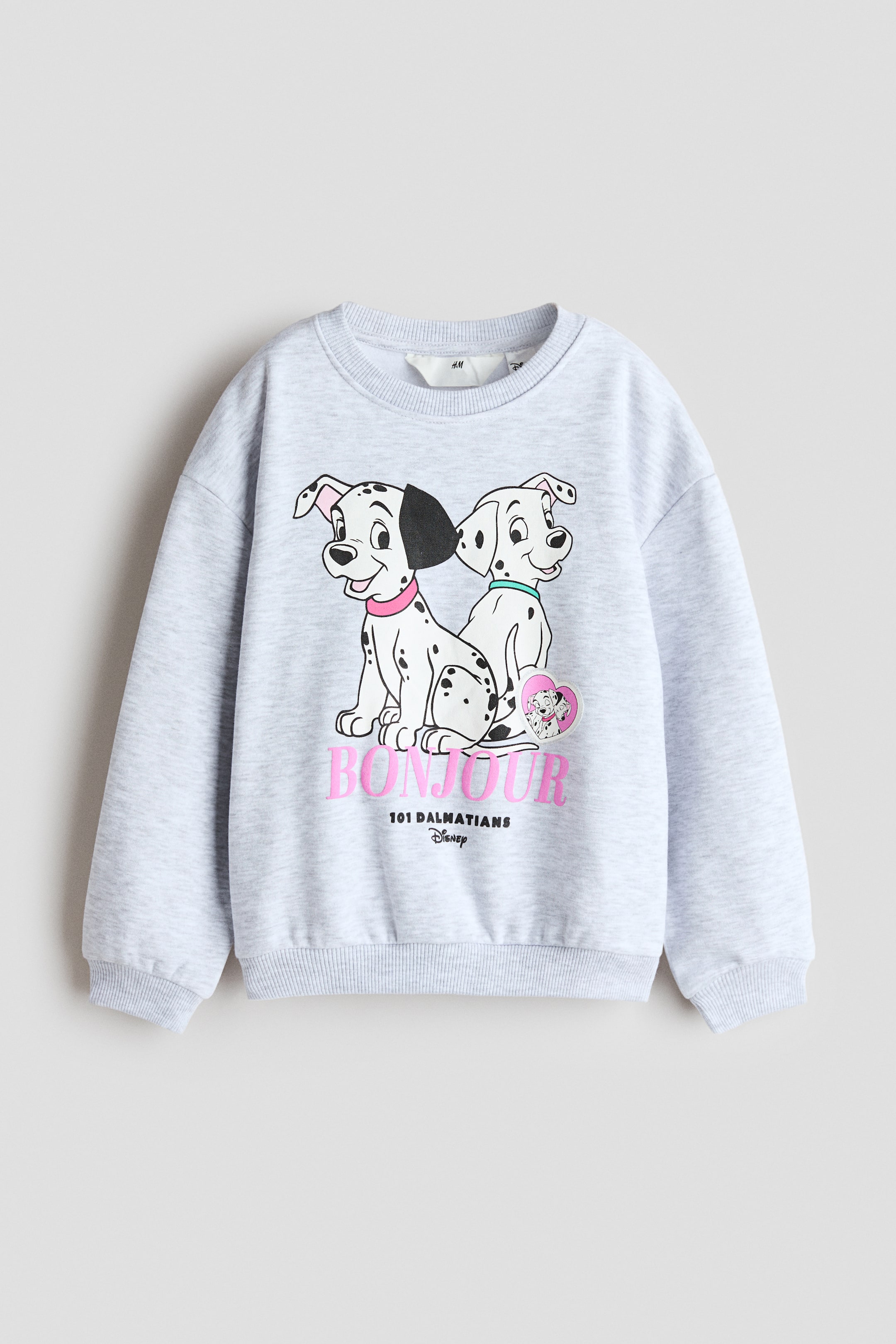 Größeres Bild anzeigen: Ein hellgrau meliertes Sweatshirt ist von vorne zu sehen, mit Rundhalsausschnitt, überschnittenen Schultern und langen Ärmeln mit Rippbündchen. Auf der Vorderseite ist ein gedrucktes Motiv von zwei Cartoon-Dalmatinern über dem Wort „BONJOUR“ in pinker Schrift zu sehen, abgerundet durch einen Rippbund.