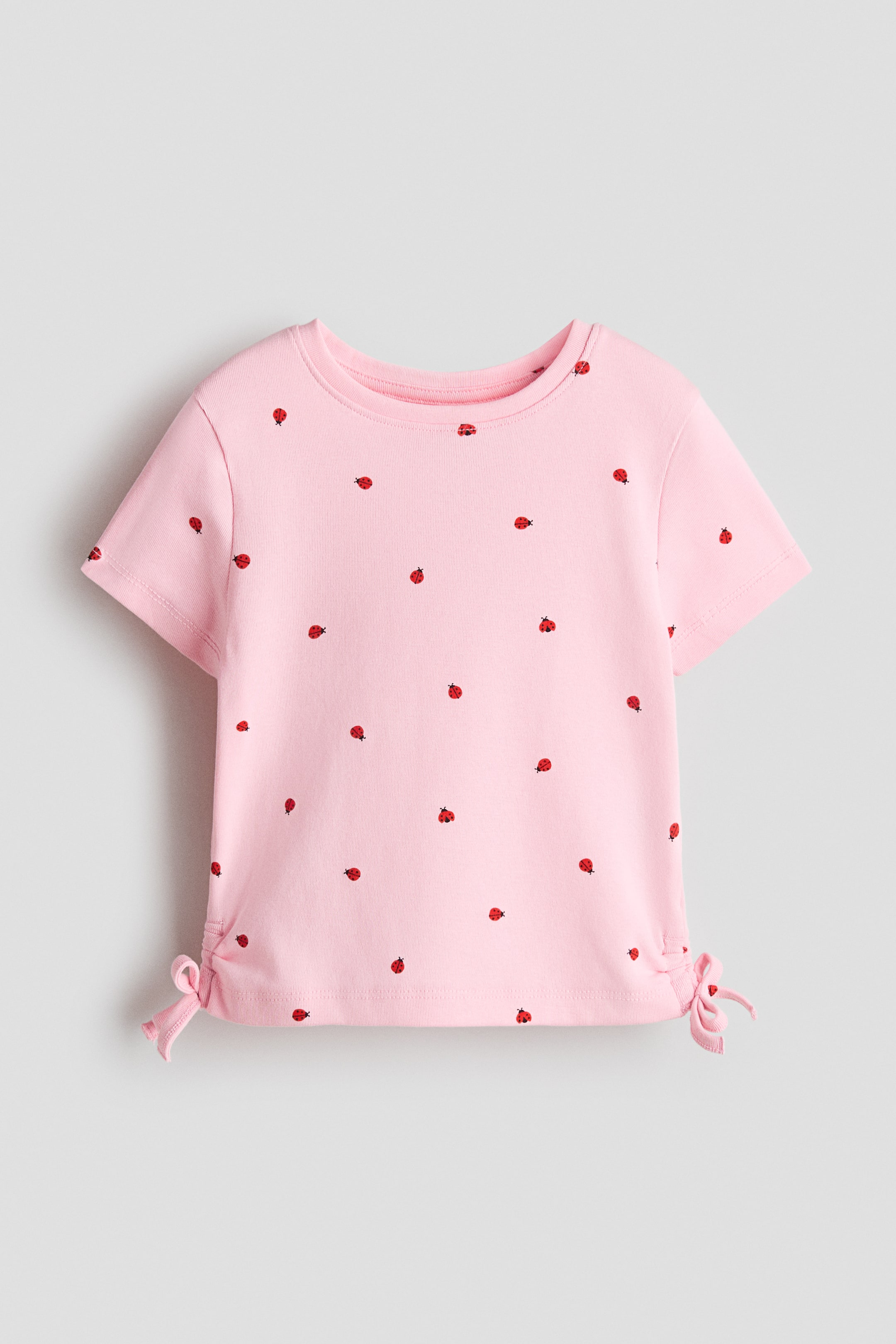 View larger image: Pink t-shirt is featuring a crew neck, short sleeves, and an all-over pattern of small red and black ladybugs. The front of the t-shirt is showcasing adjustable ties on both side seams, cinching the hem.