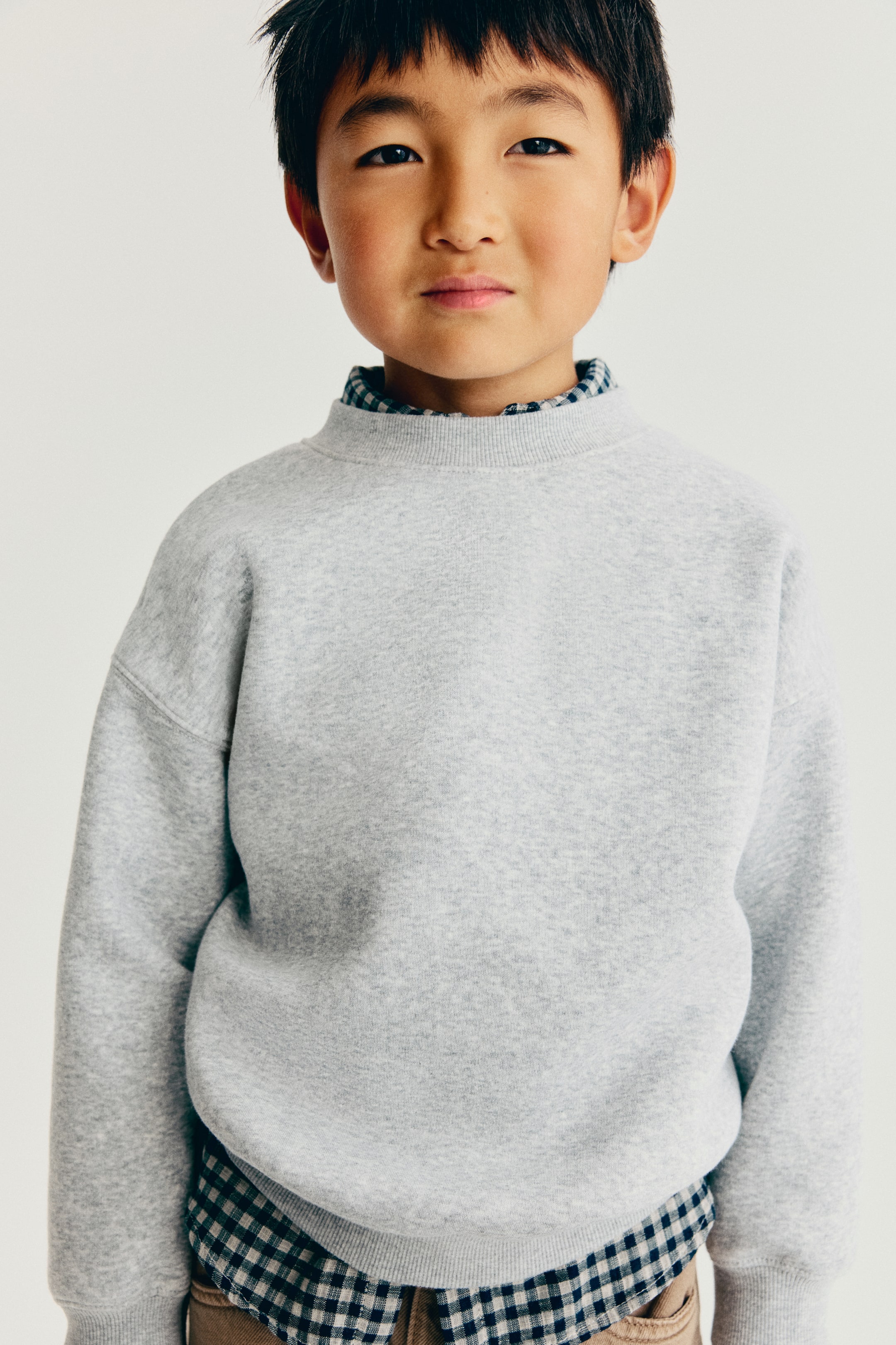 Agrandir l'image: Un sweat-shirt chiné gris clair est présenté de face, avec un col rond côtelé, des épaules tombantes, ainsi que des poignets et un ourlet côtelés. Le sweat-shirt est porté en superposition sur une chemise à carreaux bleus et blancs, dont le col et l’ourlet sont visibles.