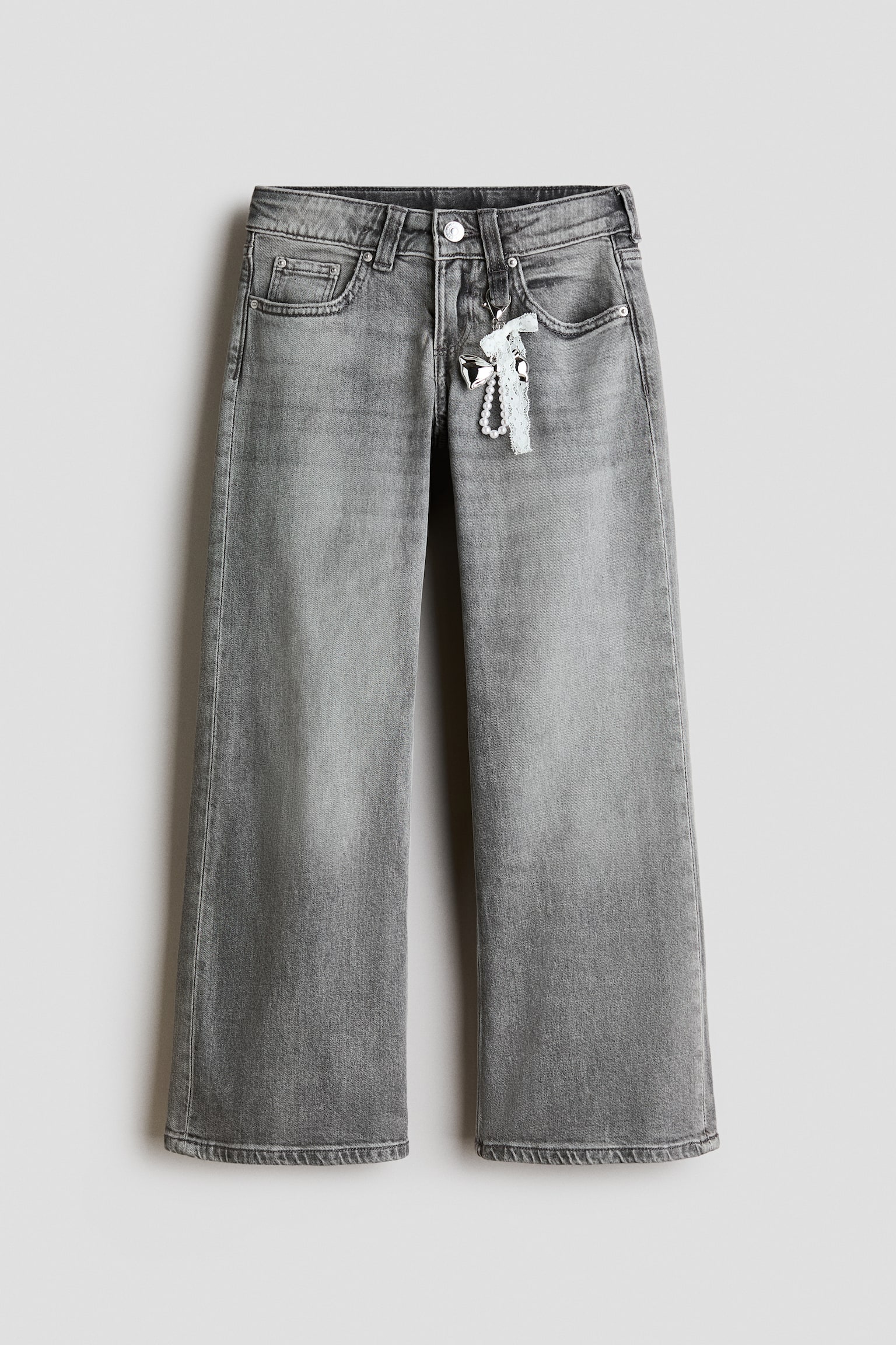 A pair of grey washed denim jeans is shown from the front, featuring a wide-leg silhouette, five-pocket styling, and a button fastening at the waist. A decorative charm featuring a white lace bow, pearl beads, and a silver heart is hanging from one of the front belt loops.