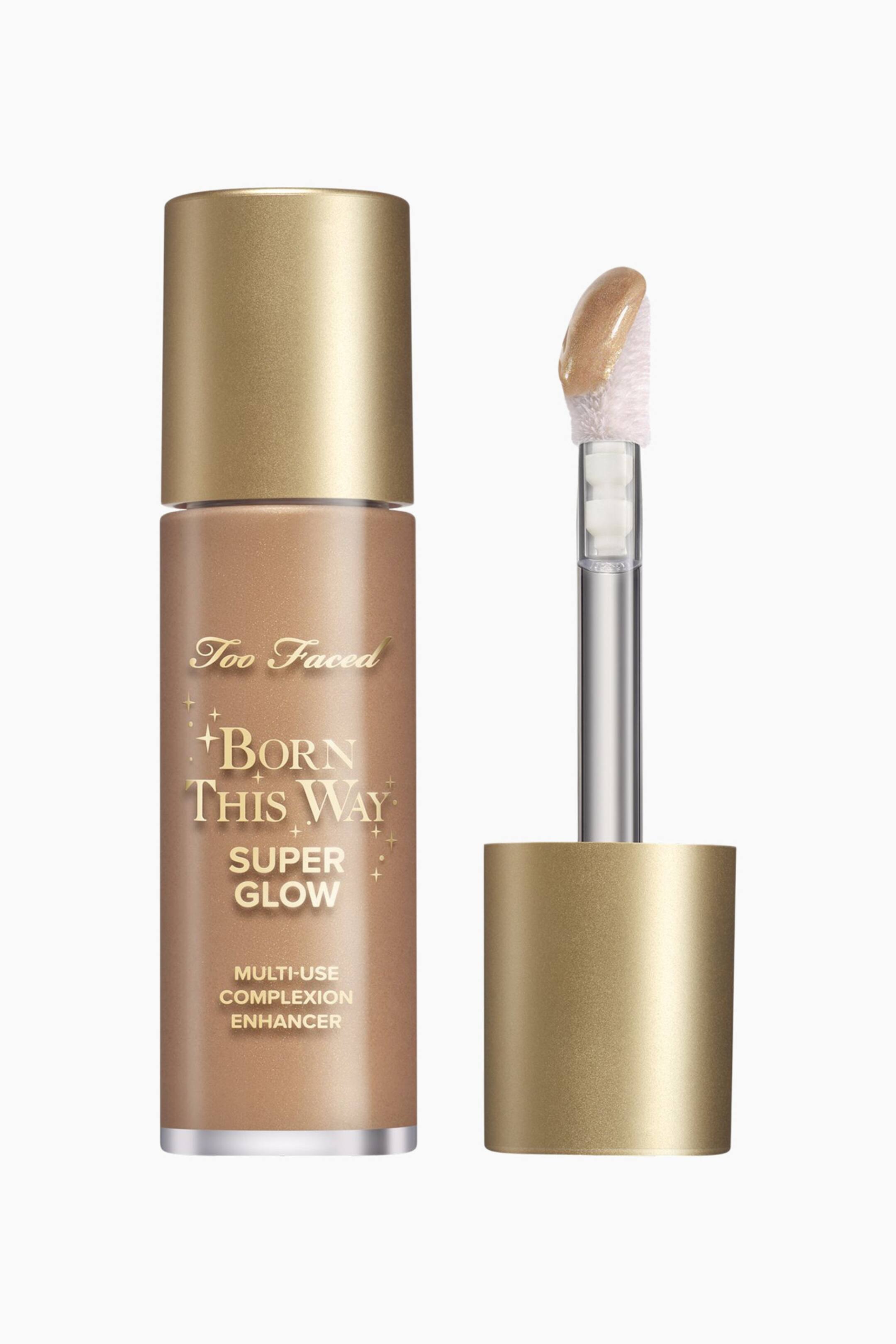 Visa större bild: En cylindrisk flaska av Too Faced Born This Way Super Glow Multi-Use Complexion Enhancer står upprätt och visar en metallisk brons-guld vätska och en guldfärgad kork. Till höger om den står produktens applikator med fotspets också upprätt, dess spets täckt med den skimrande, guld-bronsiga förstärkaren.