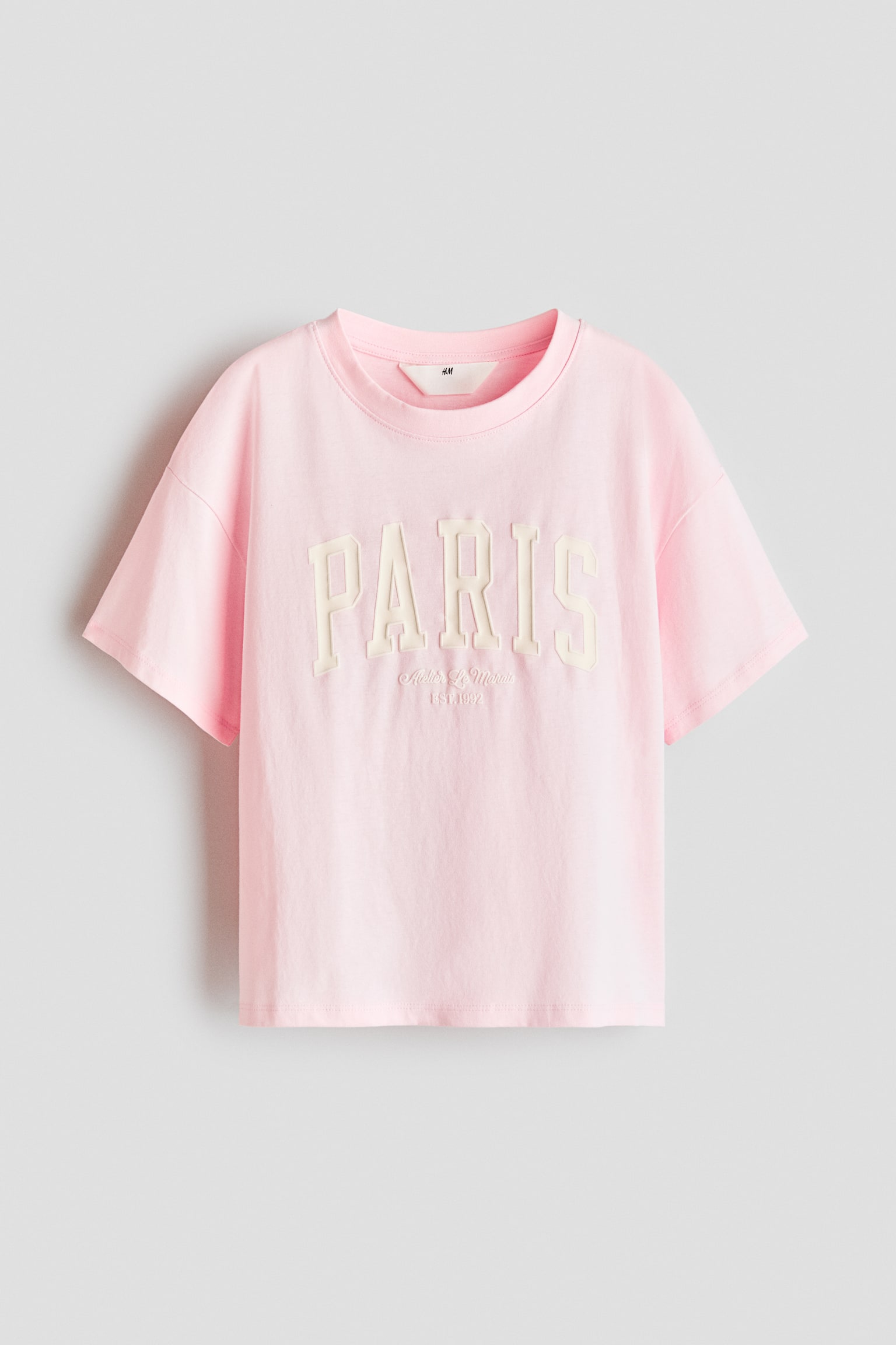 A light pink cotton jersey t-shirt is shown from the front, featuring a round rib-trimmed neckline and dropped shoulders. The chest displays a printed design featuring the word "PARIS" in off-white varsity-style block letters with smaller text reading "Atelier Le Marais EST. 1992" underneath.