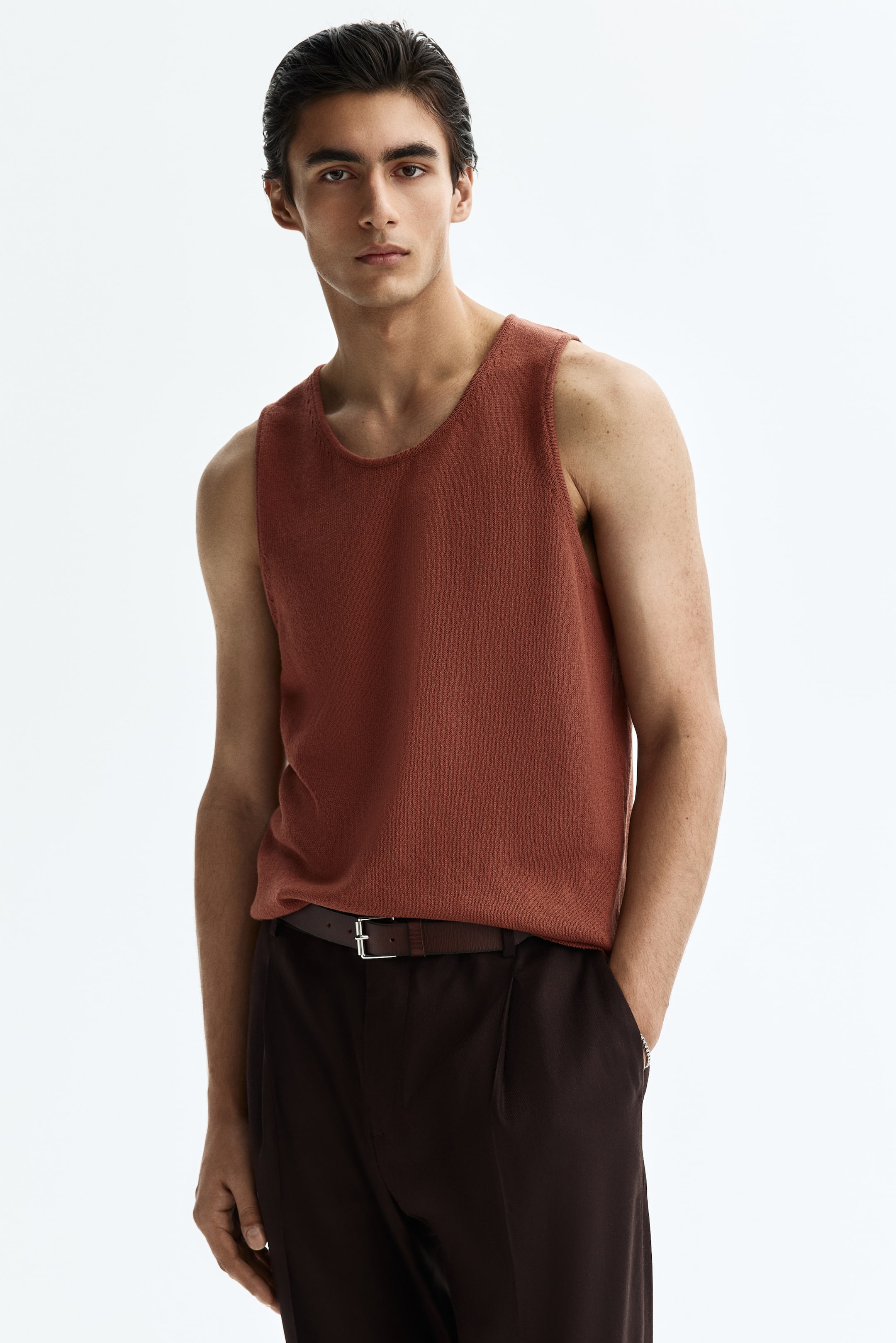 View larger image: A rust-colored knit tank top features a round neckline and wide armholes, styled partially tucked into dark brown pants.