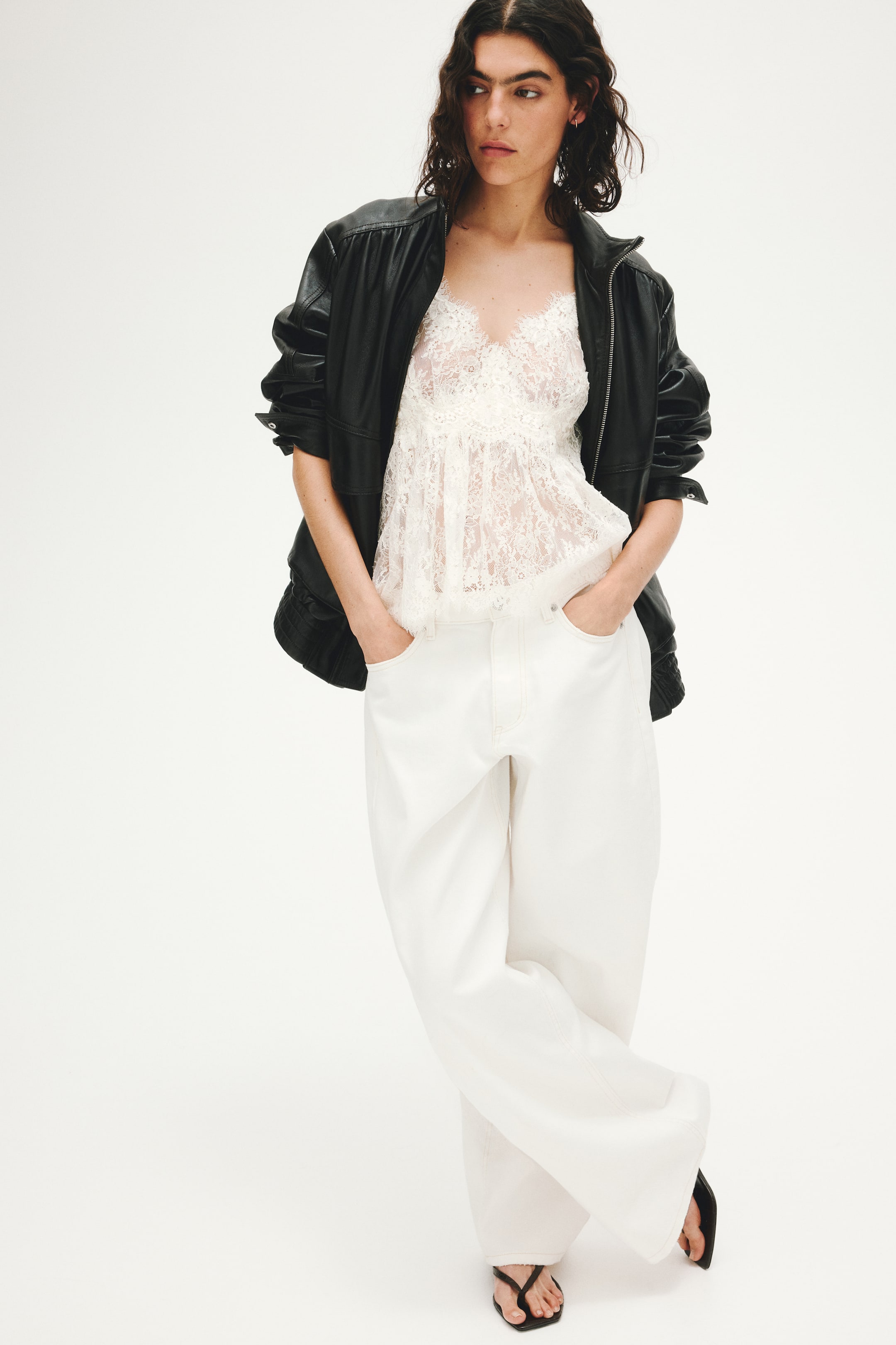 View larger image: An oversized black leather jacket, with sleeves casually pushed up, is showcased from a front-angled perspective, worn open over a white semi-sheer lace camisole that features a V-neckline. This ensemble is paired with white wide-leg trousers, highlighting visible contrast stitching, and complemented by black thong sandals.