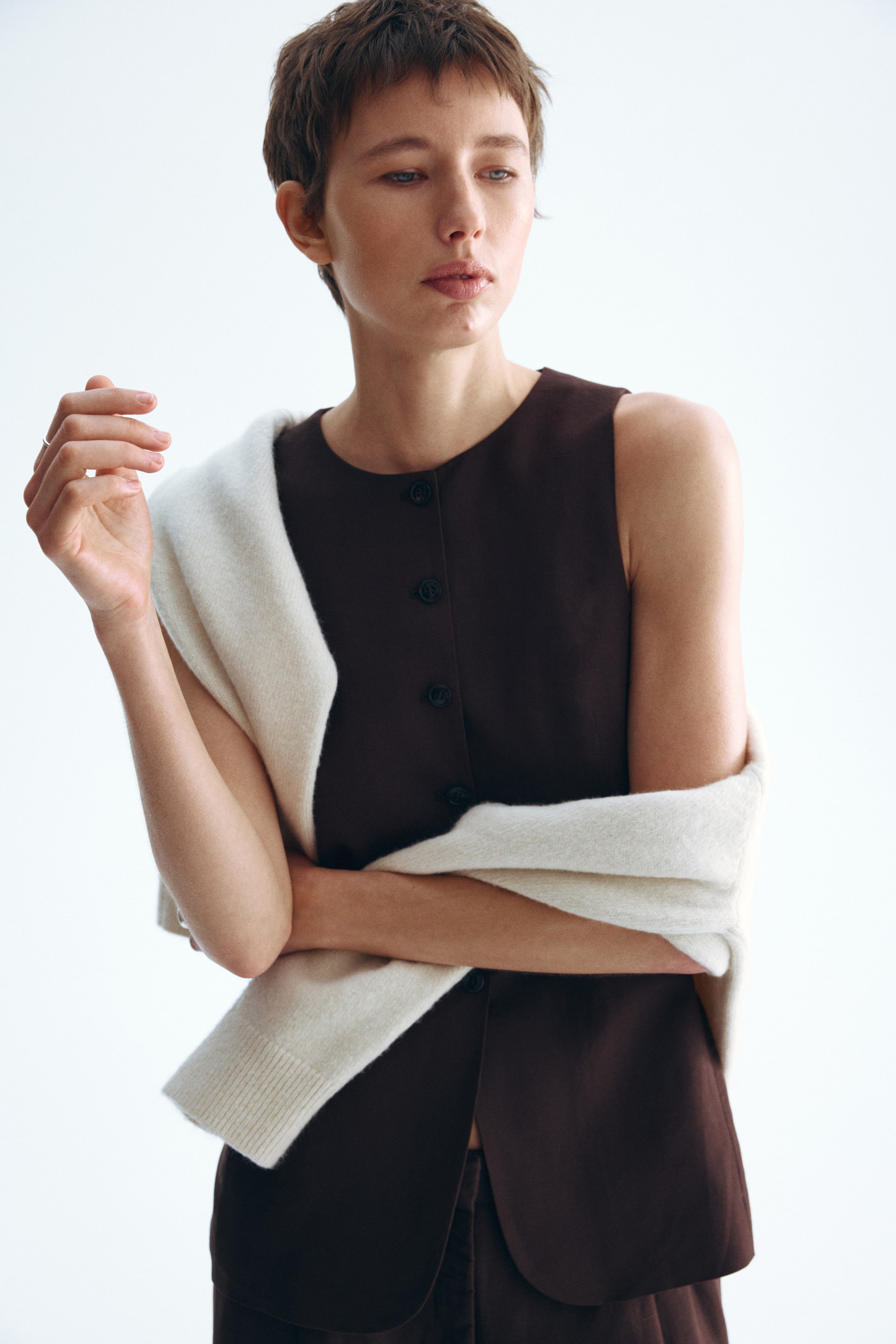 查看大圖: 深棕色無袖上衣，圓領設計，配有暗色鈕扣的鈕扣前襟。一件淺色毛衣，帶有明顯的羅紋袖口，披在肩膀和手臂上，部分覆蓋上衣。