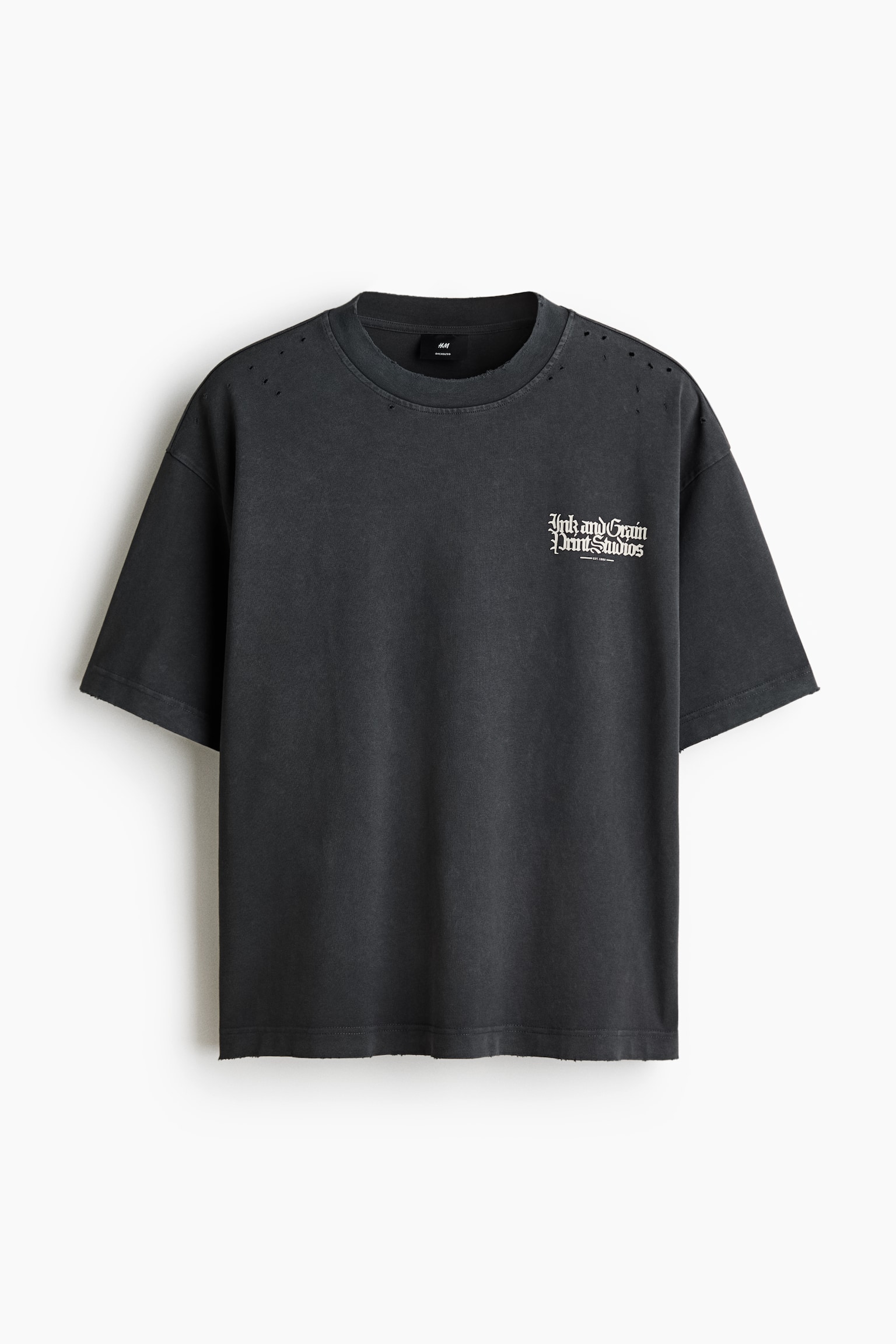 En mÃ¸rkegrÃ¥, oversized T-shirt er vist forfra og fremhÃ¦ver en lÃ¸s, baggy silhuet med sÃ¦nkede skuldre og brede Ã¦rmer. Den viser distressed-detaljer, herunder smÃ¥ huller nÃ¦r den ribbede rundhals og skuldrene, et hvidt gotisk tekstprint pÃ¥ venstre bryst og en diskret frynset kant.
