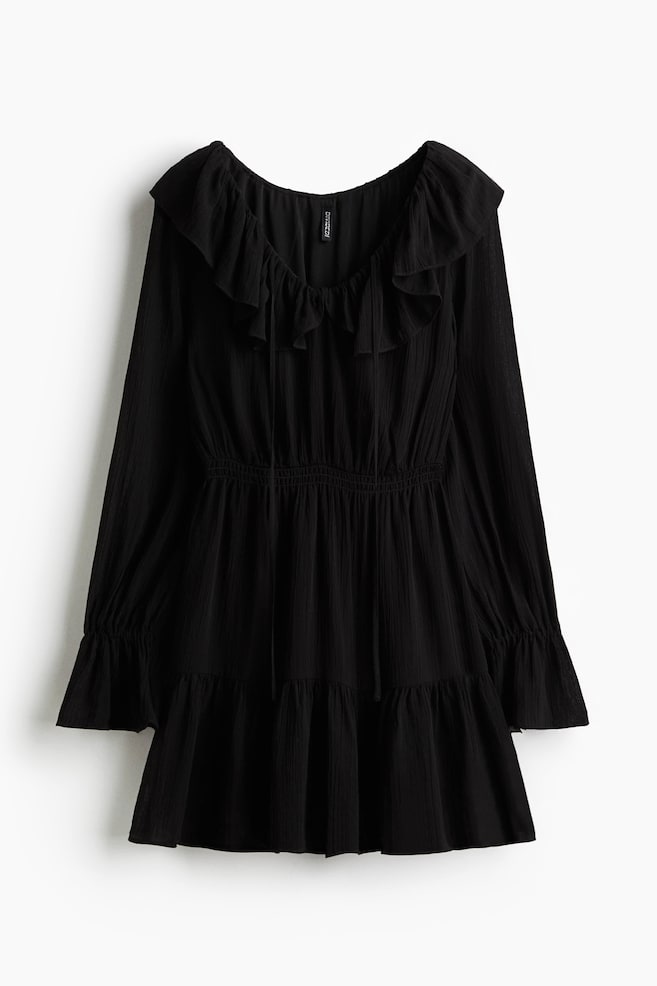 Un vestido negro arrugado presenta un escote redondo con un volante ancho superpuesto y lazos frontales. Está diseñado con mangas largas que terminan en puños elásticos, una cintura fruncida y una falda con volantes. - 1