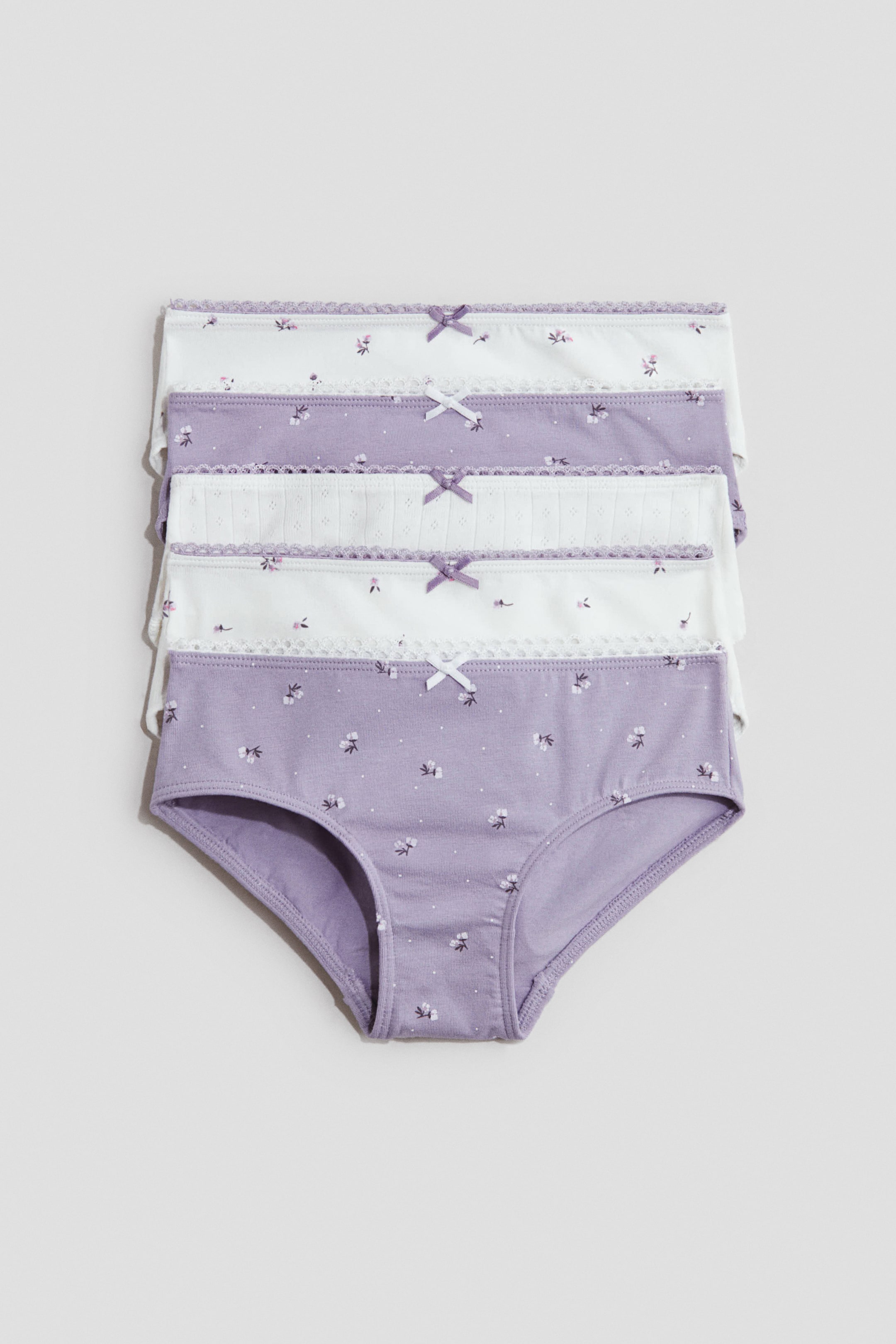 Grotere afbeelding bekijken: Vijf slips zijn gestapeld en tonen zacht katoenen jersey-stof. Elke slip heeft een picot-afgewerkte tailleband met een decoratieve satijnen strik aan de voorkant en een gevoerde kruisje, terwijl ze worden gepresenteerd in afwisselende ontwerpen van wit met paarse bloemenpatronen, paars met witte bloemenpatronen, en wit met een geribbelde textuur.