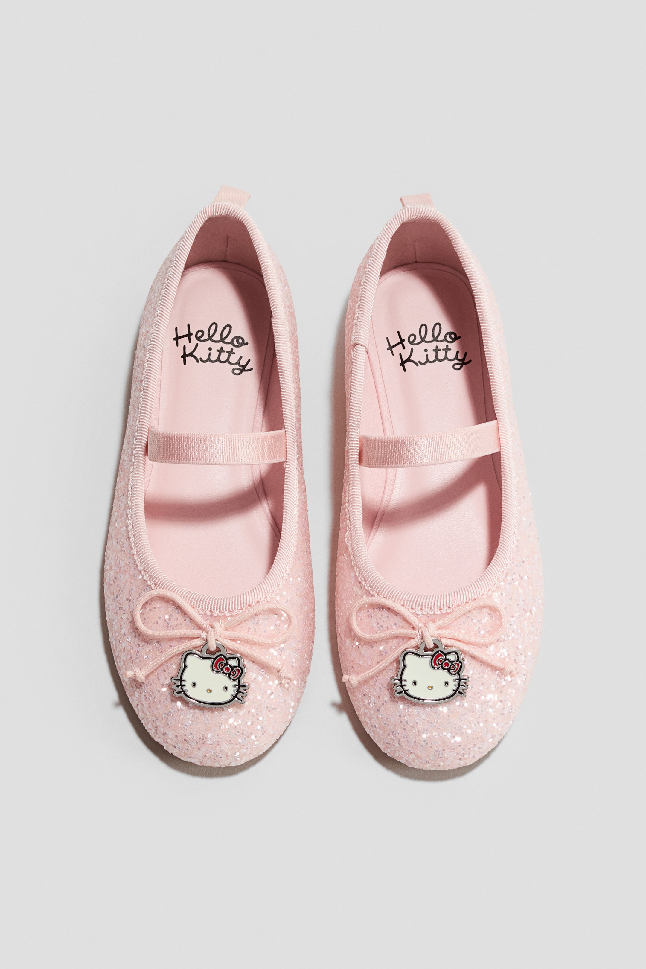 Ampliar la imagen: Un par de zapatillas de ballet rosas con brillo está orientado hacia ADELANTE, mostrando correas elásticas sobre el pie, moños decorativos en la puntera con dijes colgantes de la cabeza de Hello Kitty y la marca 'Hello Kitty' visible en las plantillas.