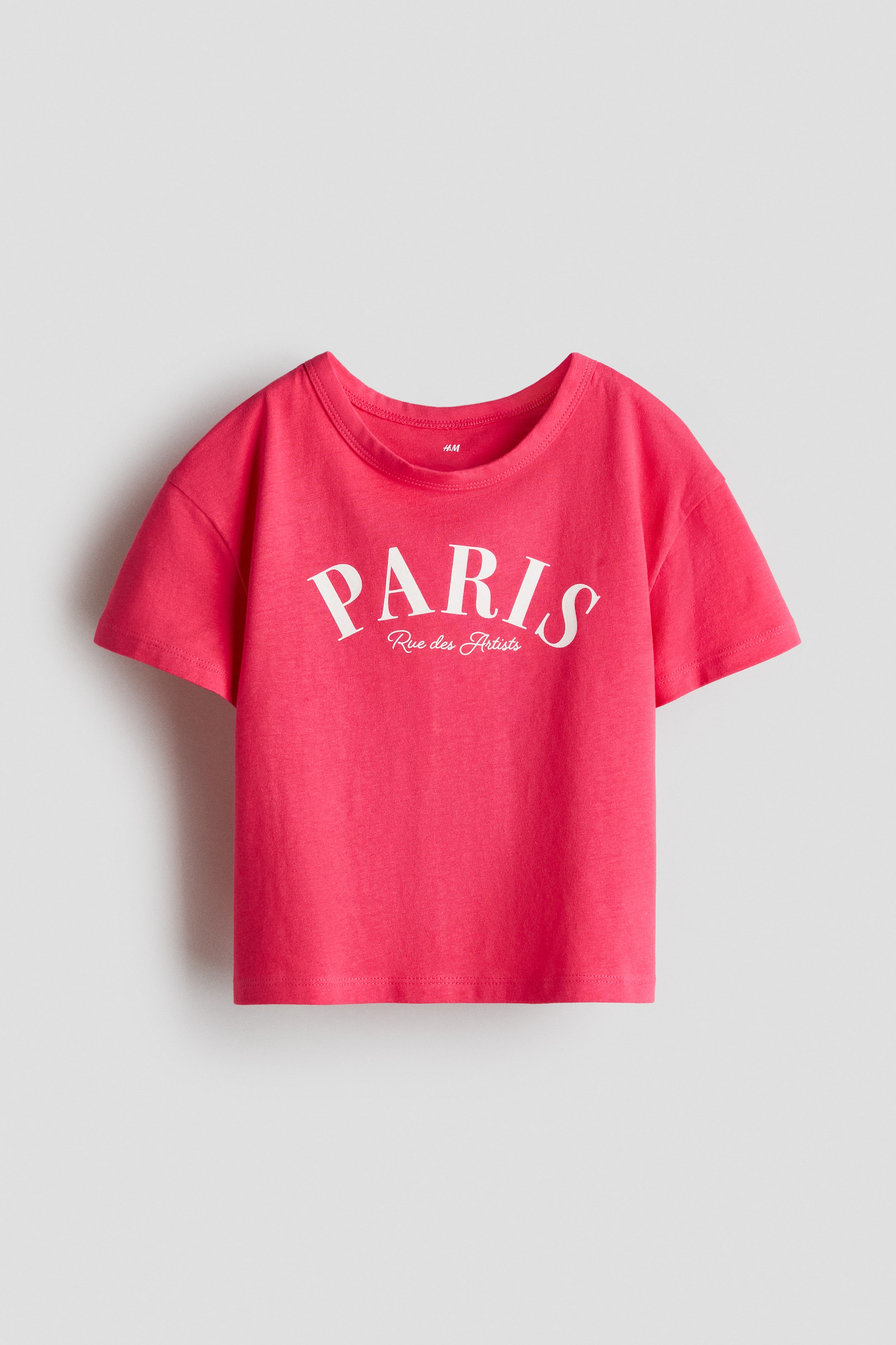 View larger image: A pink t-shirt is facing front, featuring a round neck and short sleeves. It is displaying "PARIS" in large white script font with "Rue des Artists" in smaller script below.