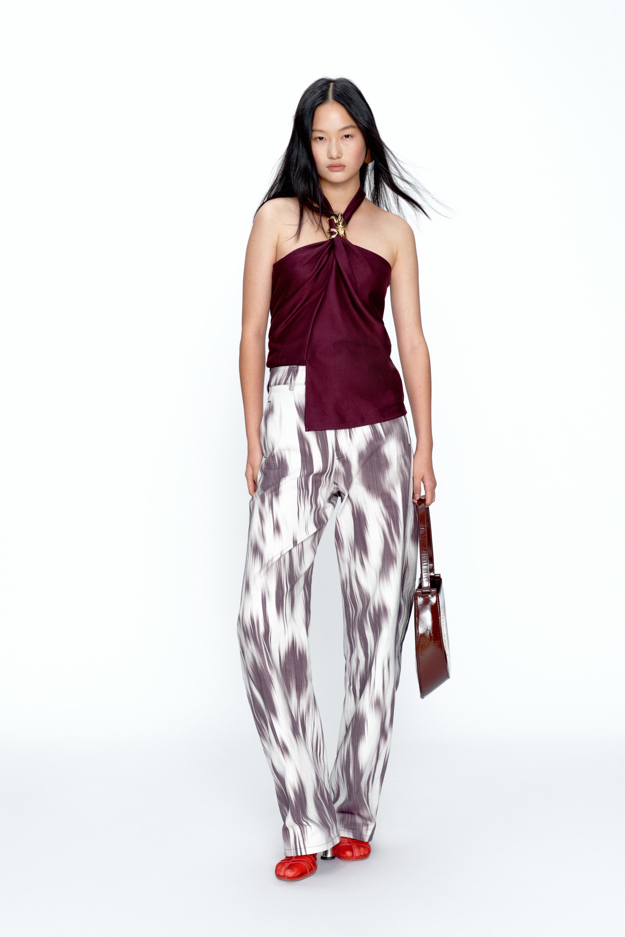 View larger image: A burgundy halter top with a gold-tone ornate buckle is styled with wide-leg trousers. The trousers feature an abstract grey and white pattern and an asymmetrical wrap detail at the waist. Red gathered flats and a glossy deep reddish-brown shoulder bag complete the look.