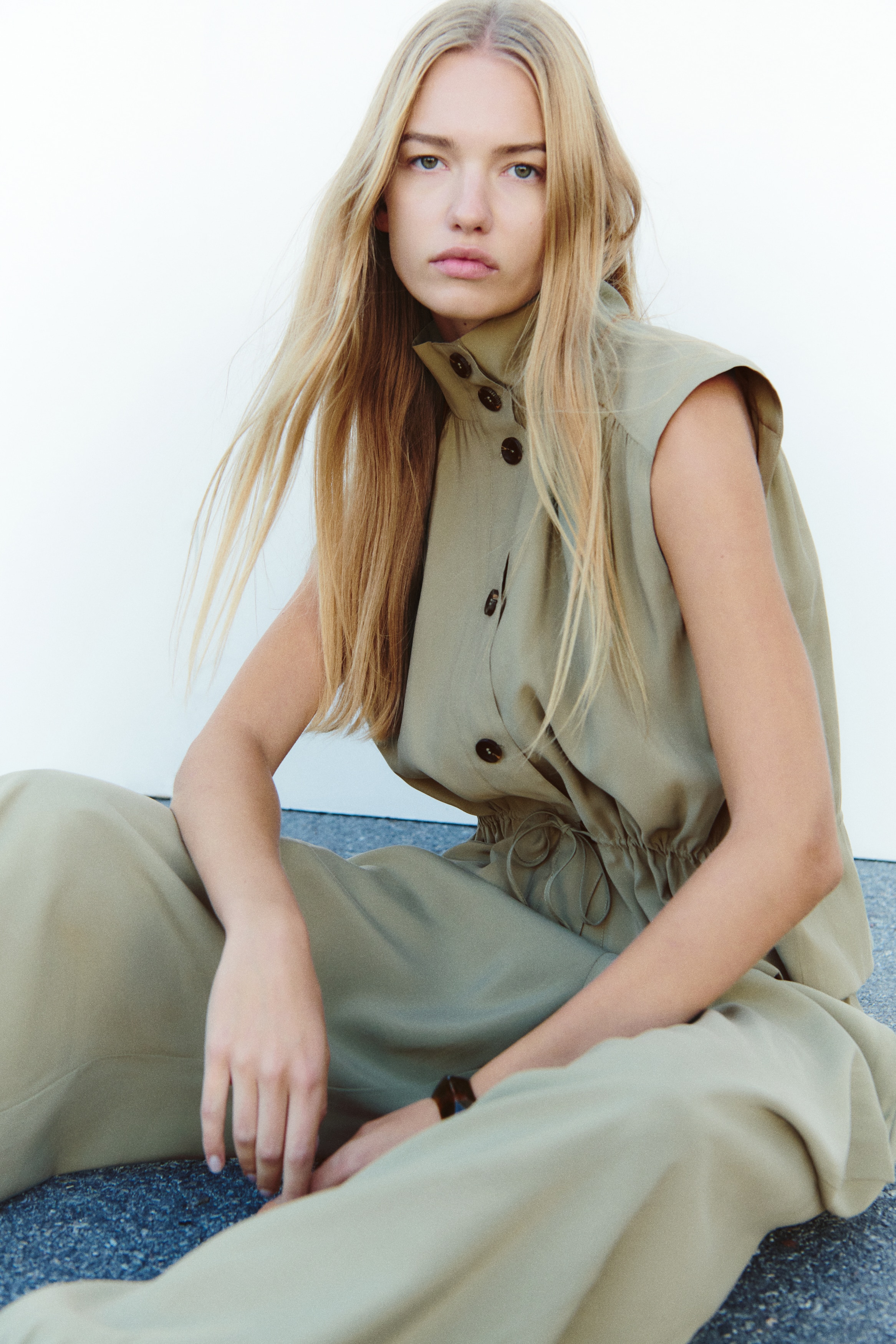 A khaki sleeveless top is facing forward, featuring a high stand-up collar and a button-front placket with four dark brown buttons. It is detailed with gathered fabric across the shoulders and upper bodice, and an elasticized drawstring waist is tied in a knot.