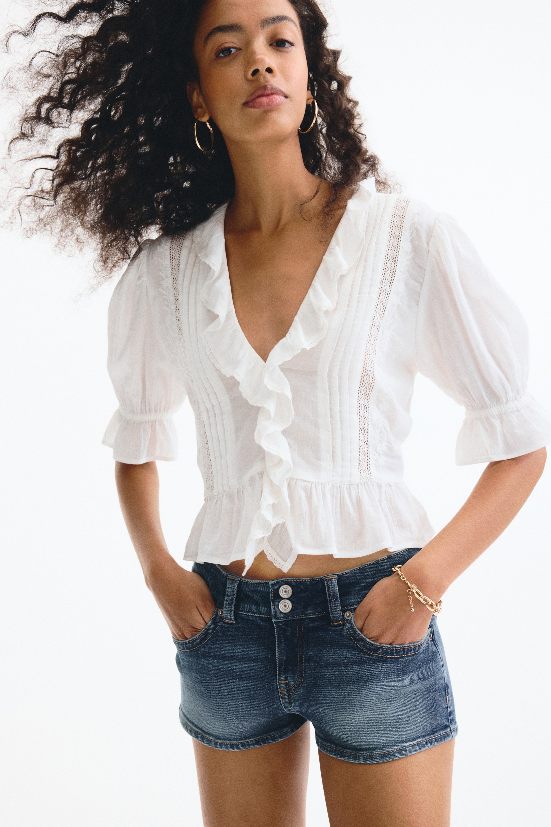 Grösseres Bild ansehen: Eine weisse Peplum-Bluse mit einem gerafften V-Ausschnitt, Biesen und kurzen Puffärmeln mit elastischen Bündchen. Dieses Oberteil wird mit dunkel gewaschenen Jeansshorts kombiniert, die über einen Doppelknopfverschluss und vordere Schaufeltaschen verfügen.