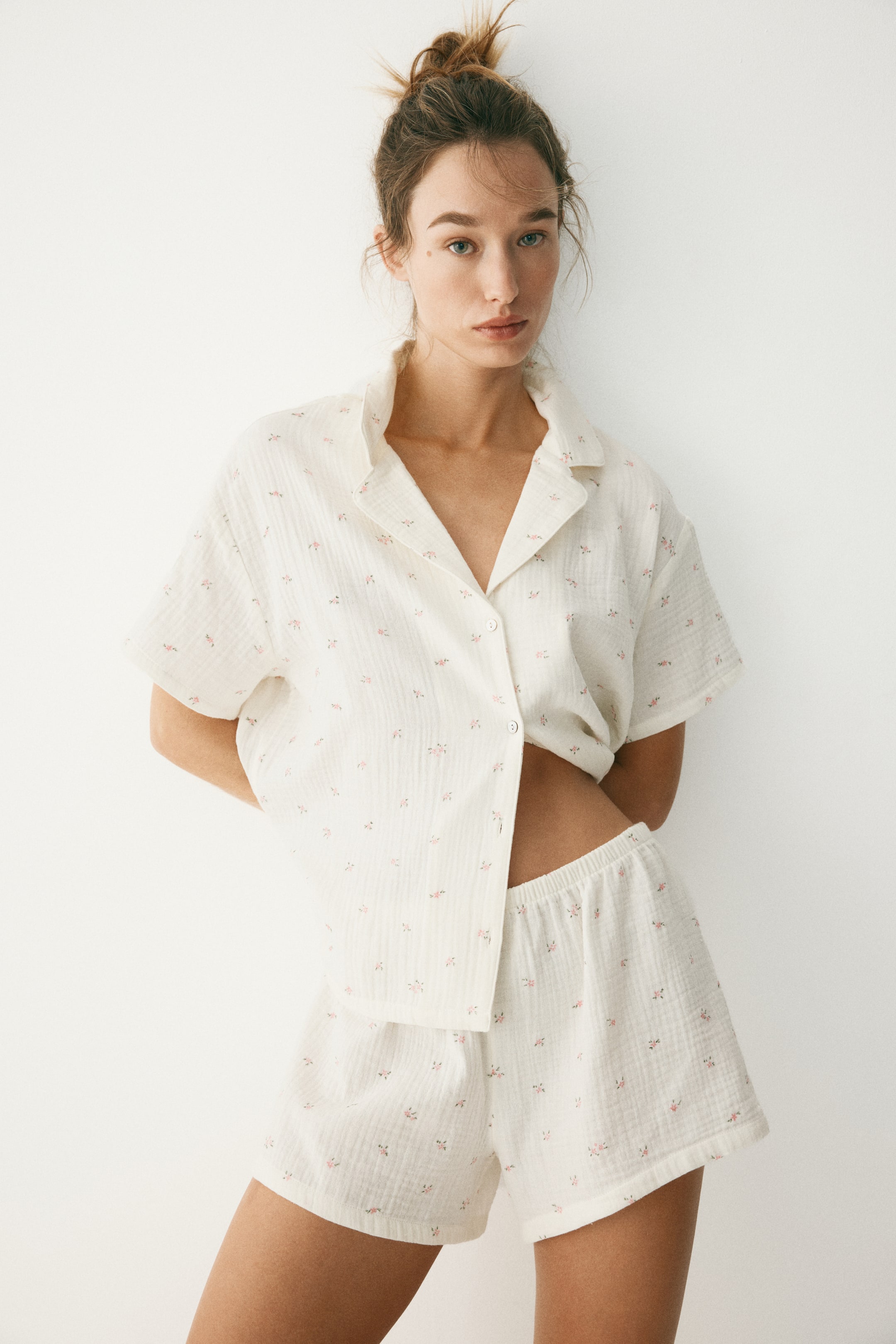 Visa större bild: Ett off-white pyjamasset, tillverkat av ett texturerat tyg, innehåller en kortärmad, krageförsedd skjorta med knappar framtill och matchande shorts, alla prydda med ett subtilt rosa blommönster. Skjortan är delvis uppknäppt nertill, vilket avslöjar midjan, och shortsen har en elastisk midja.