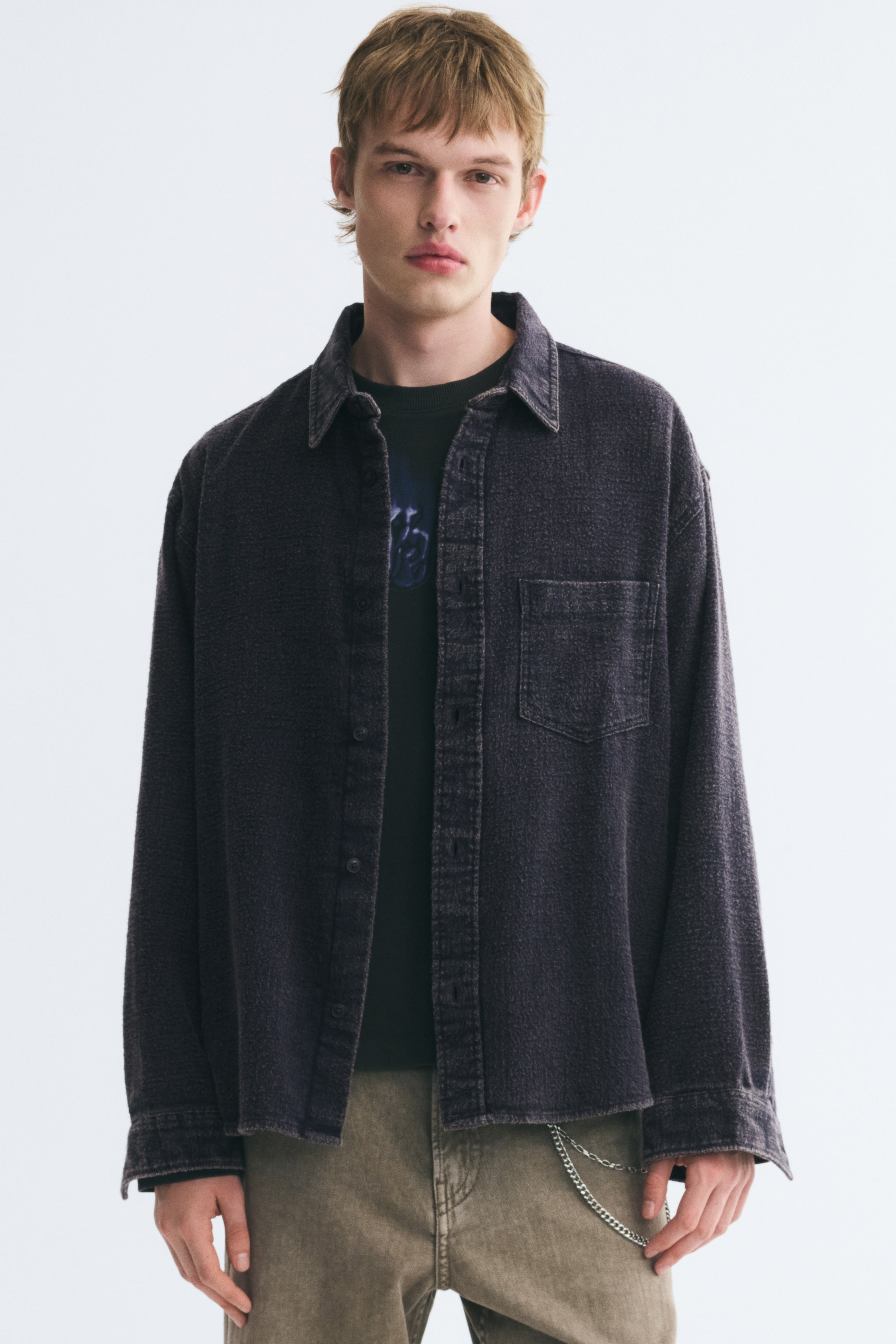 View larger image: A dark purple cotton twill overshirt with a distressed finish is worn open by the model, featuring a collar, a single chest patch pocket, and long sleeves with button cuffs. The overshirt is layered over a dark T-shirt and paired with light brown trousers.